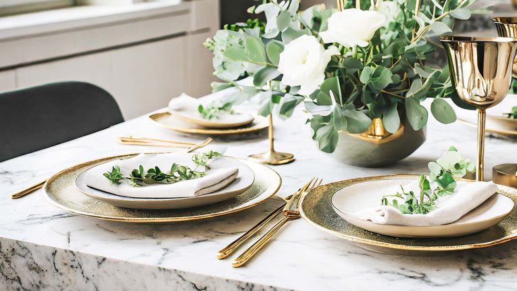 Gold flatware set stylized on marble dining table with plates, napkins, and vase filled with greenery.