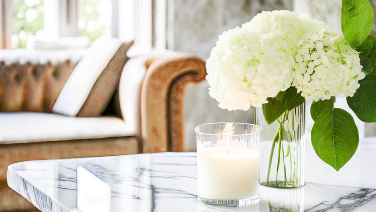 Scented soy candle on marble table with hydrangeas in glass vase and tufted leather sofa in background.