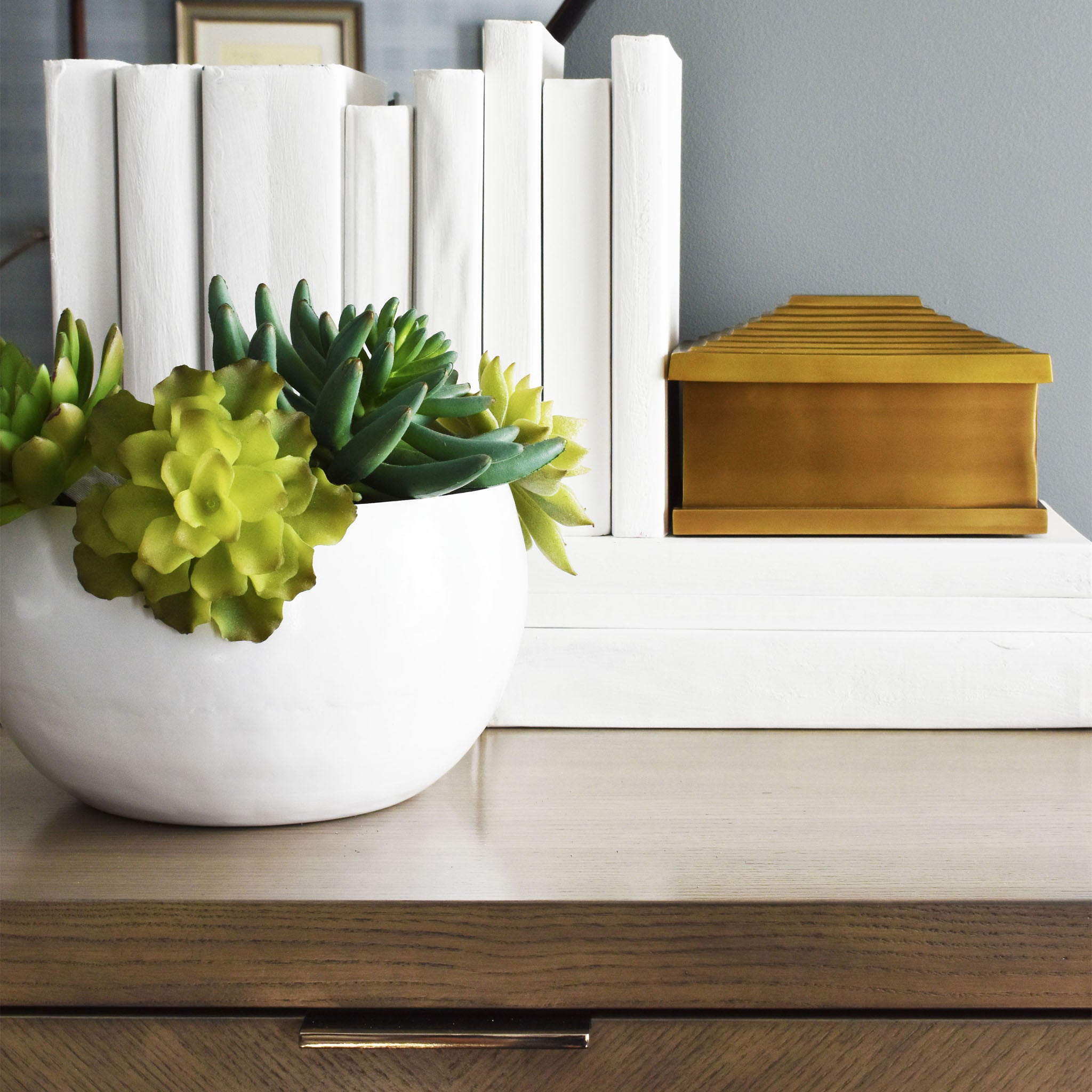 White enamel bowl with antique brass interior finish filled with succulents in front of white stacked books and brass pyramid box.