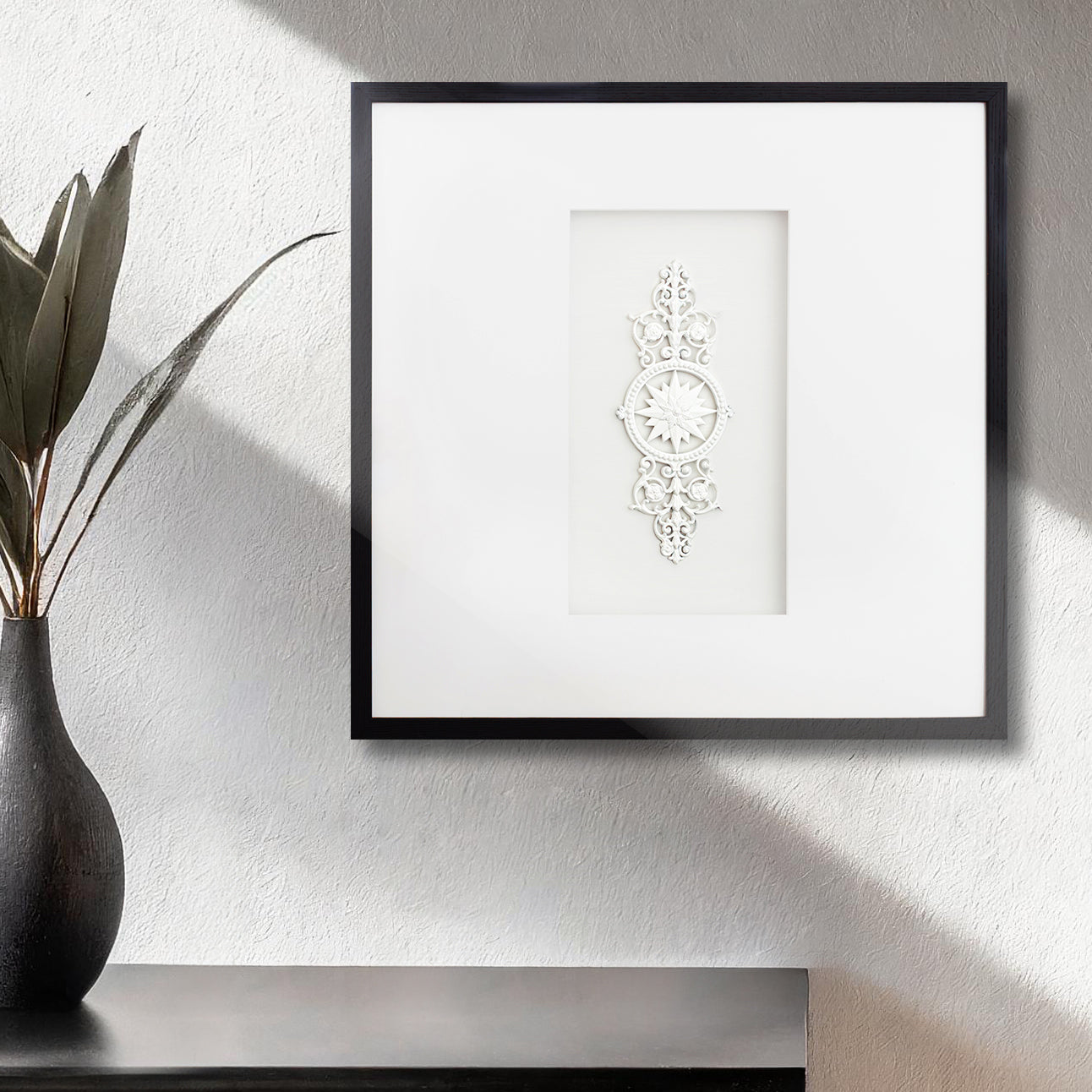 Contemporary framed sunburst rosette artwork on textured wall with vase and plant.