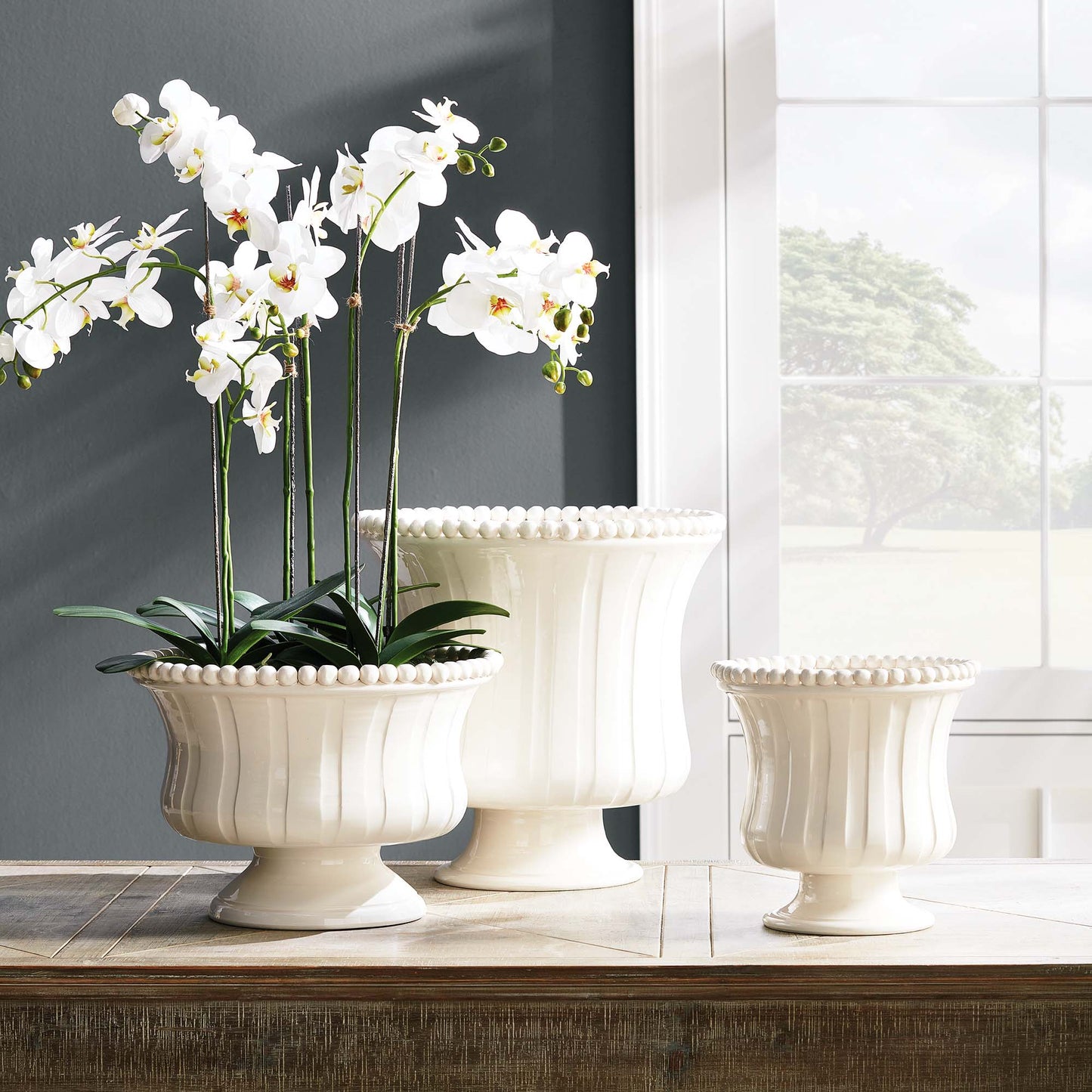 Decorative white ceramic footed bowl filled with orchid drop-in, stylized on table with white vases.