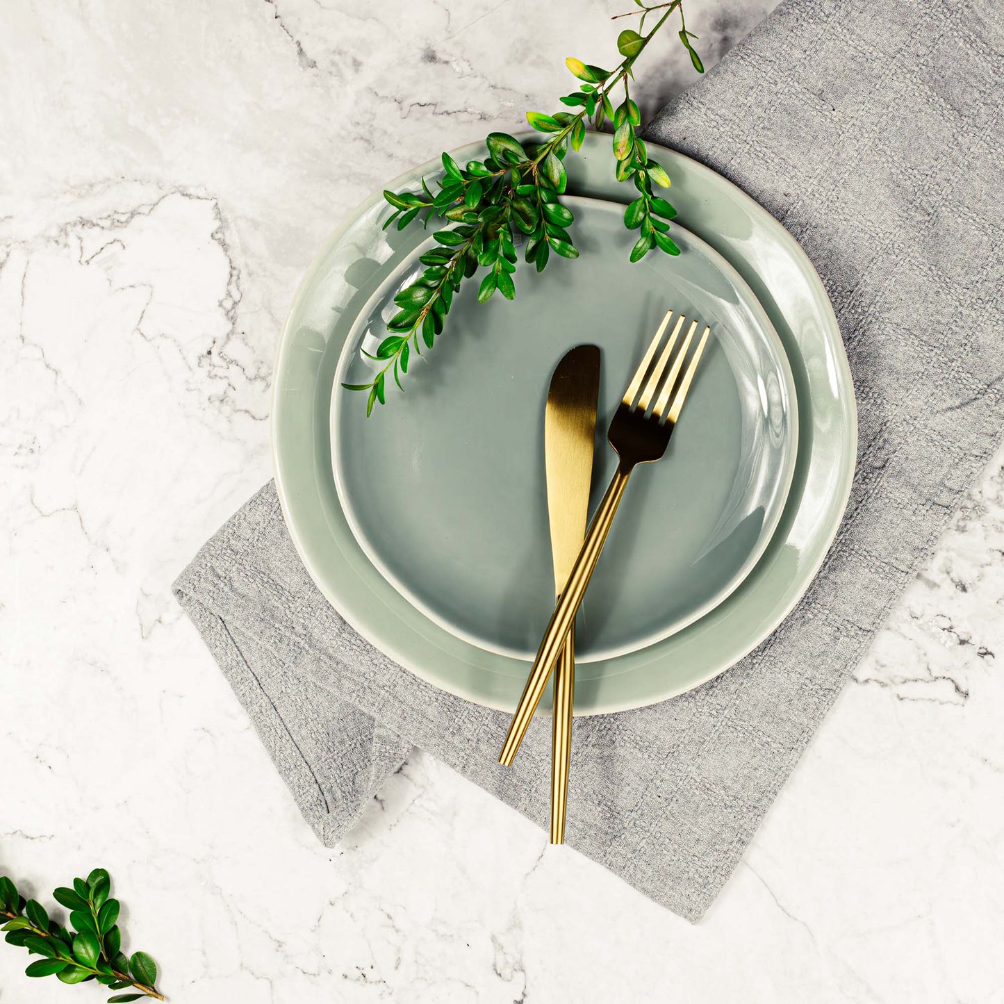 Matte gold knife and fork stylized with plates and boxwood stems on marble tabletop.