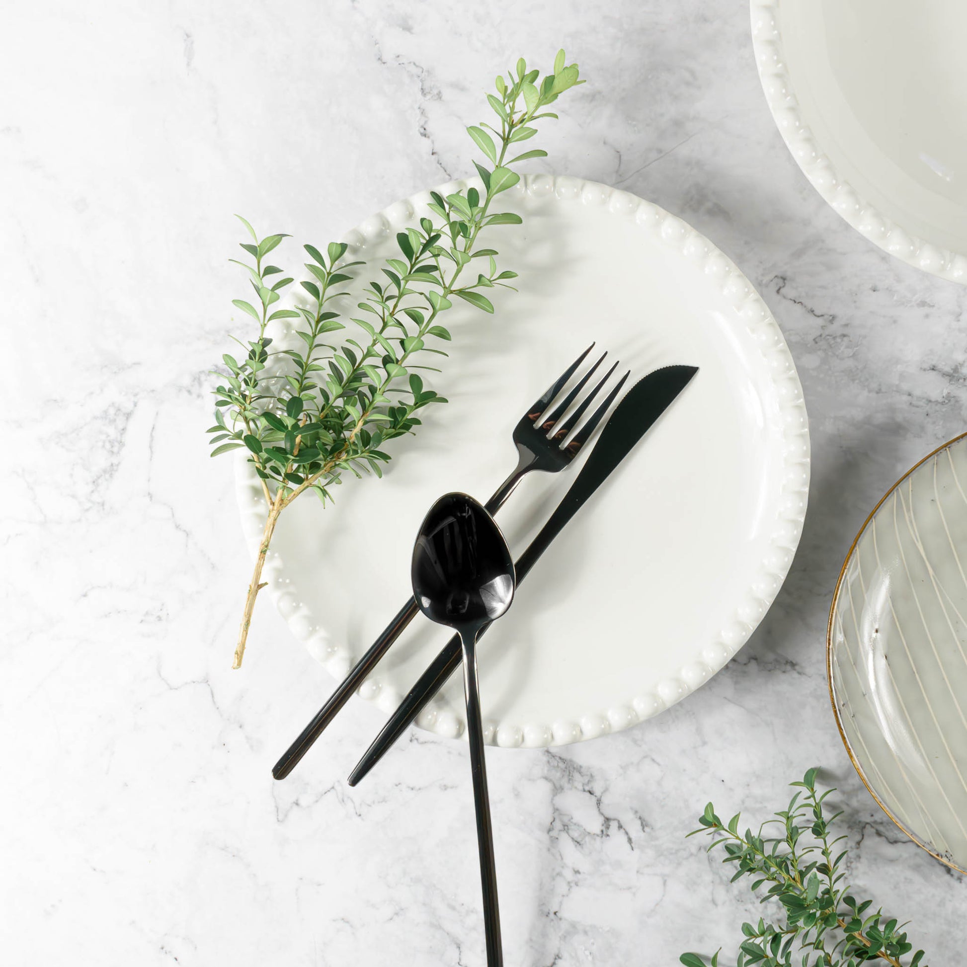 Flatware set in polished black, stylized on marble table with boxwood branches, plates, and bowls.