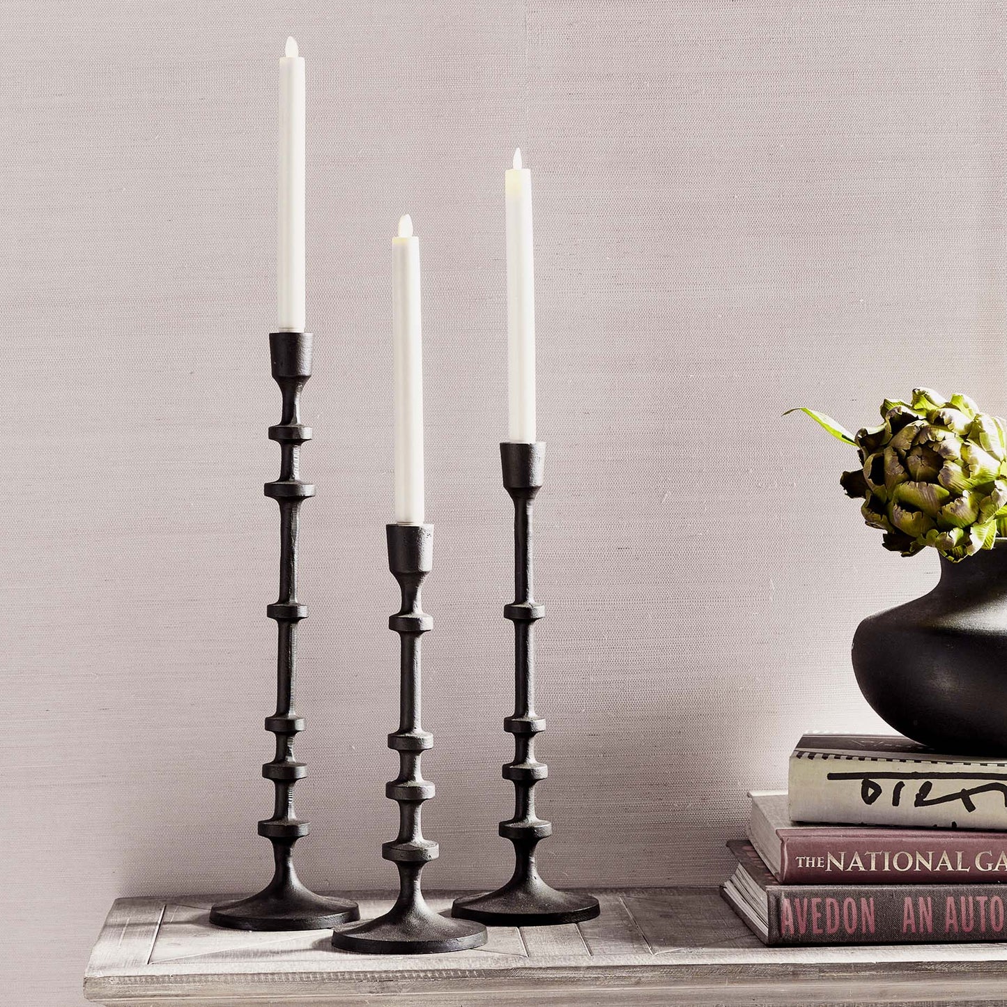Set of three minimalist taper candleholders in bronze finish stylized on console table with books, vase, and plant.