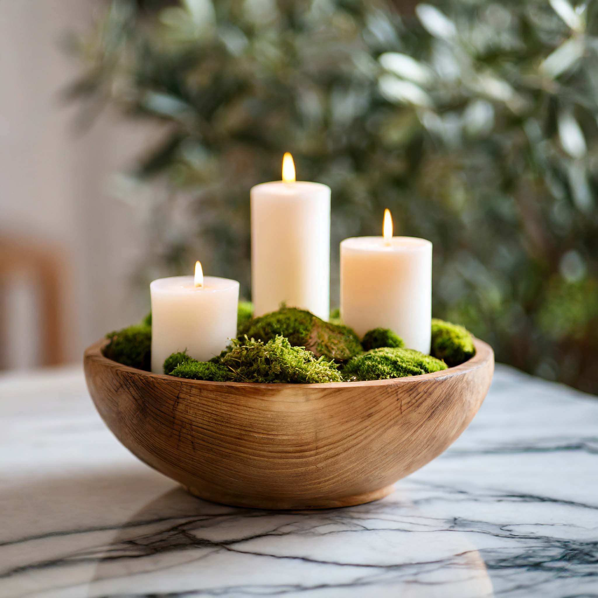 Round wooden dough bowl filled with moss and candles on marble table with olive trees in the background.