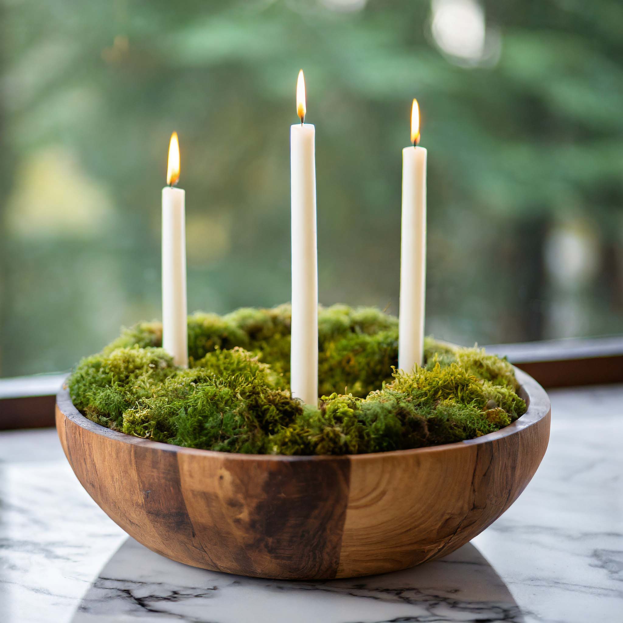 Round wooden dough bowl filled with moss and tapered candles on marble table in front of window.