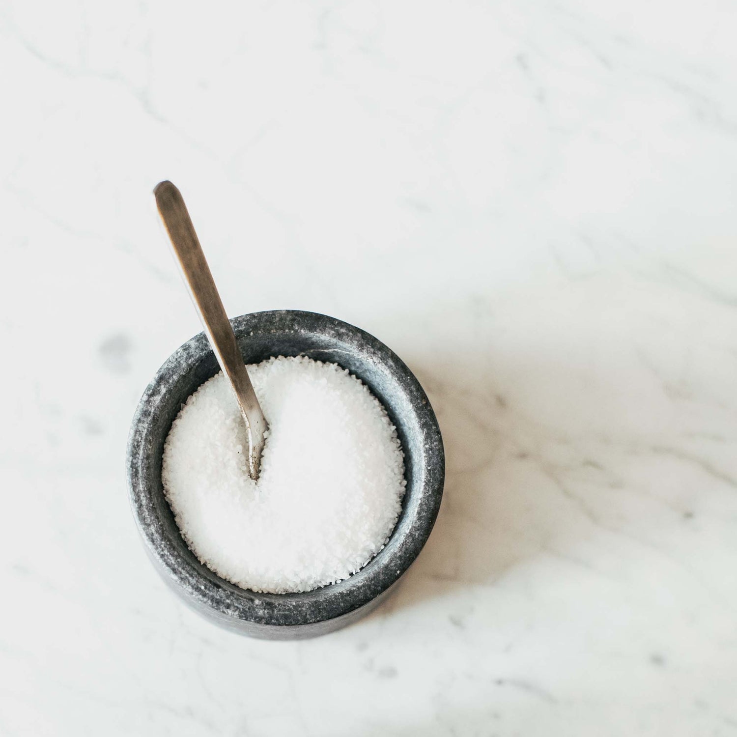 Salt in soapstone bowl with spoon on marble counter.