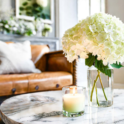 Scented soy candle on marble table with glass vase filled with hydrangeas with leather sofa in background.