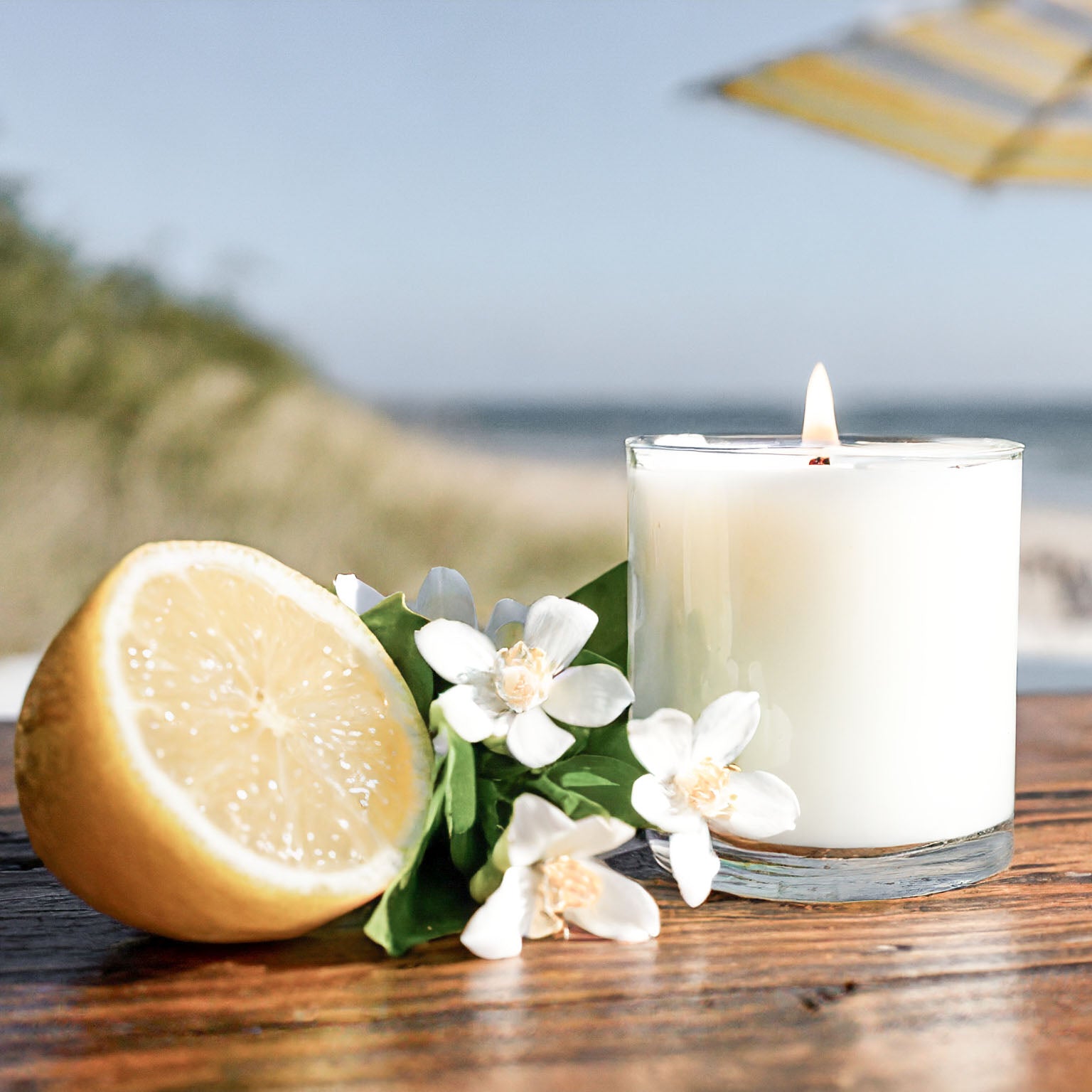 Scented soy candle on teak wood table with lemon and jasmine with sea oats, beach, and umbrella in background.