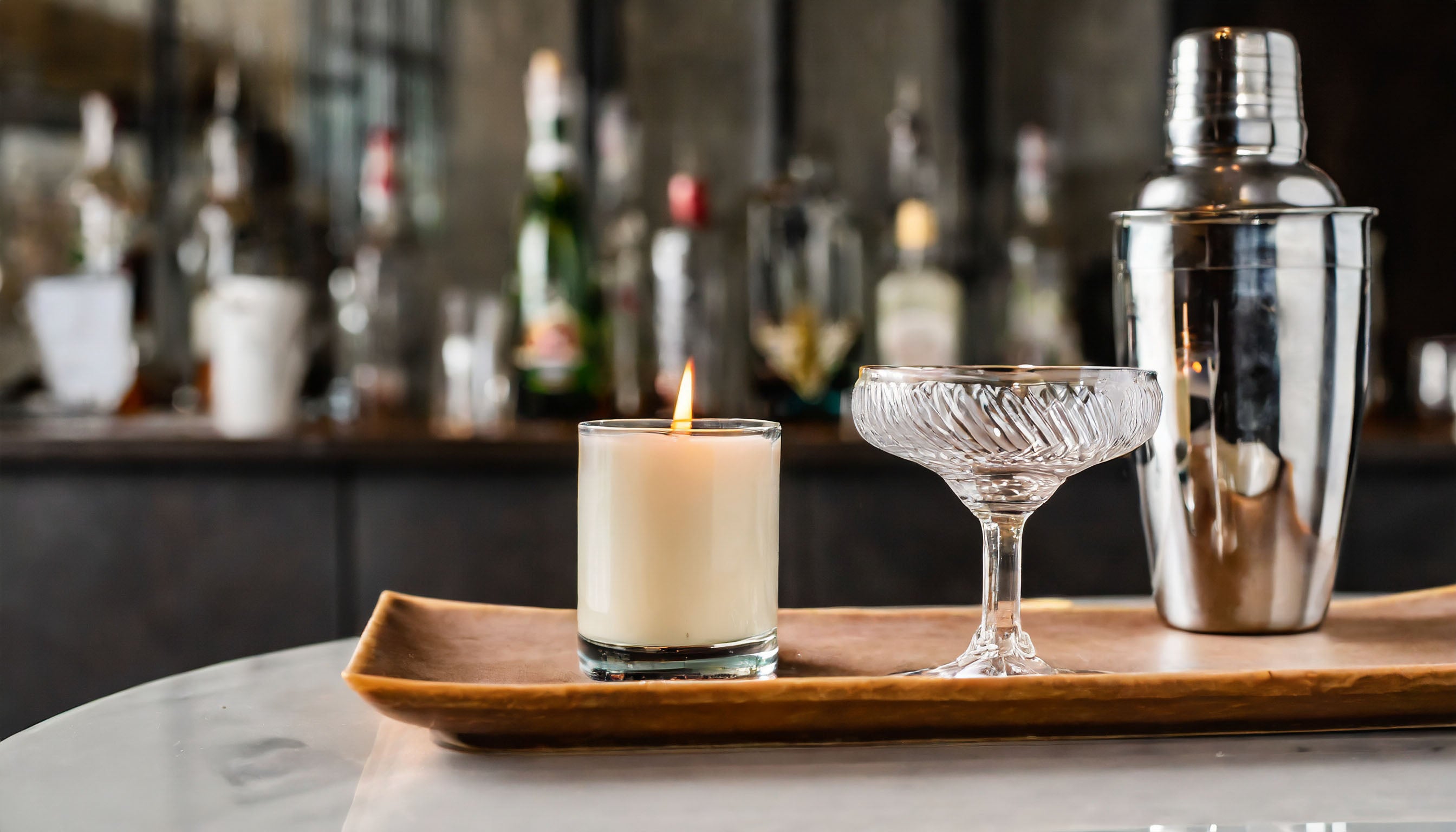 Scented soy candle styli shed on leather tray with drink glass and martini shaker with bar in background.
