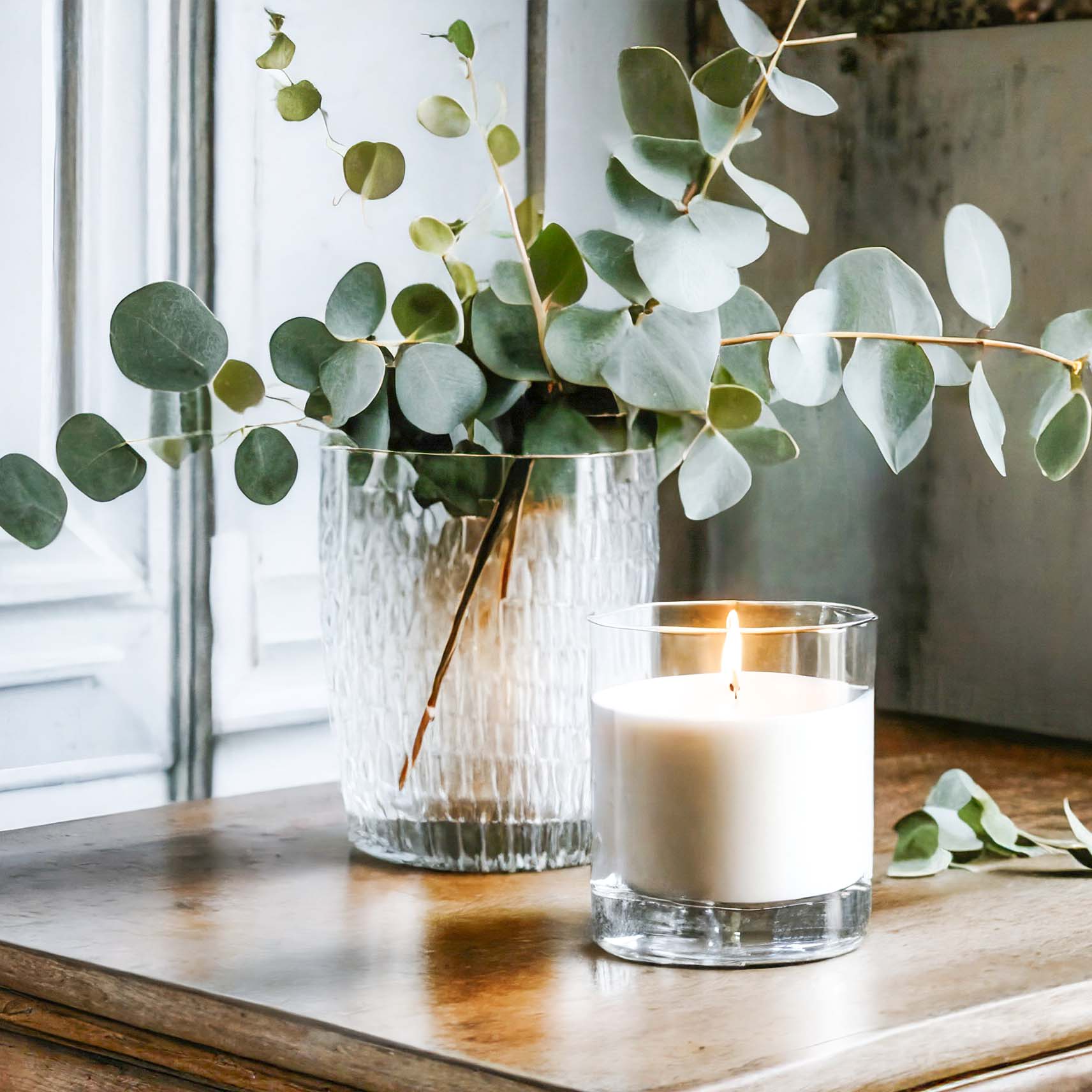 Scented soy wax blend candle on wood table with glass vase filled with eucalyptus stems.