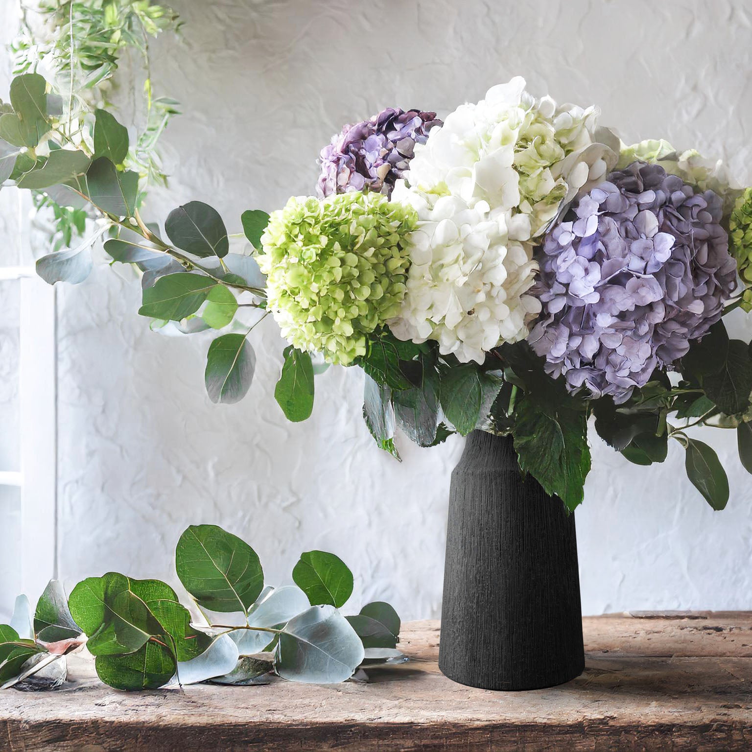 Matte black textured vase filled with flowers on wooden table in front of window.