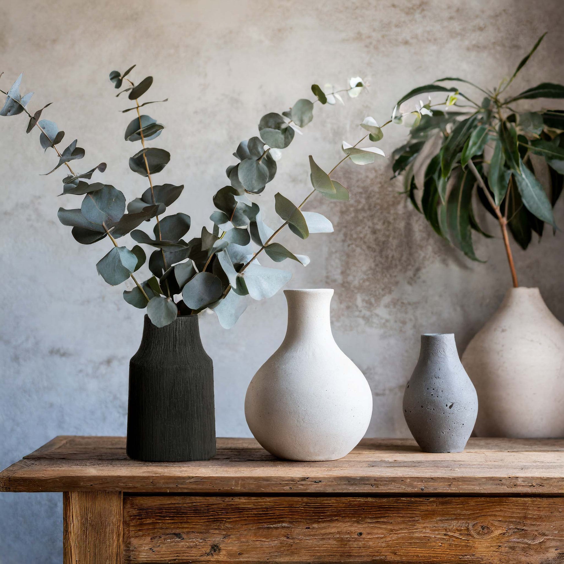 Textured ceramic small vase in matte black, filled with eucalyptus stems, on rustic wood table with ceramic vases.