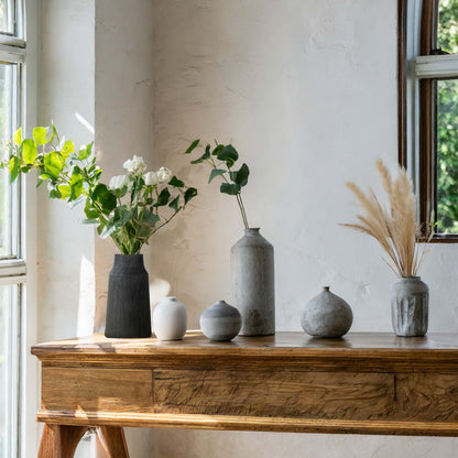Matte black textured ceramic vase with white and gray vases on wooden table with windows and sunlight.