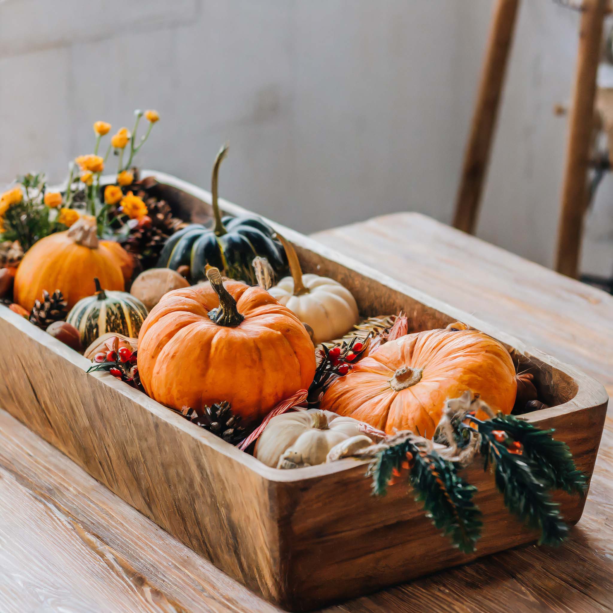 Wooden dough bowl filled with artisanal pumpkins, pinecones, flowers, and holly.
