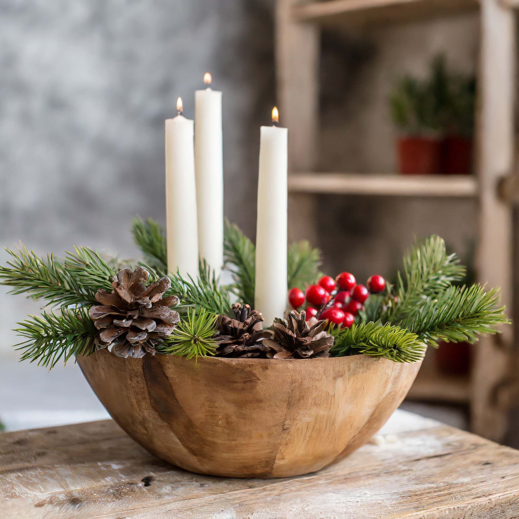 Round wooden dough bowl filled with greenery, pine cones, berries, and tapered candles.