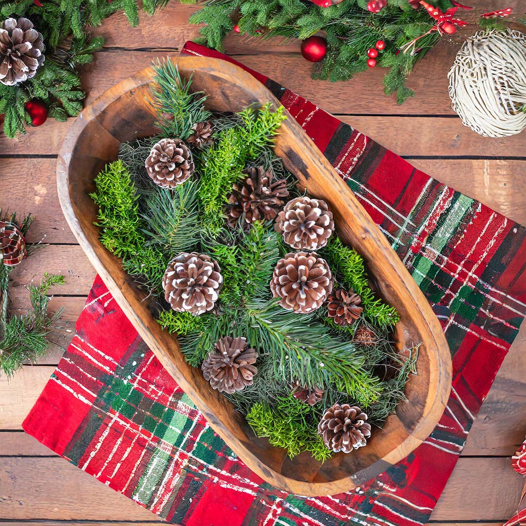 Wooden dough bowl filled with greenery and pinecones, on table with plaid tablecloth.