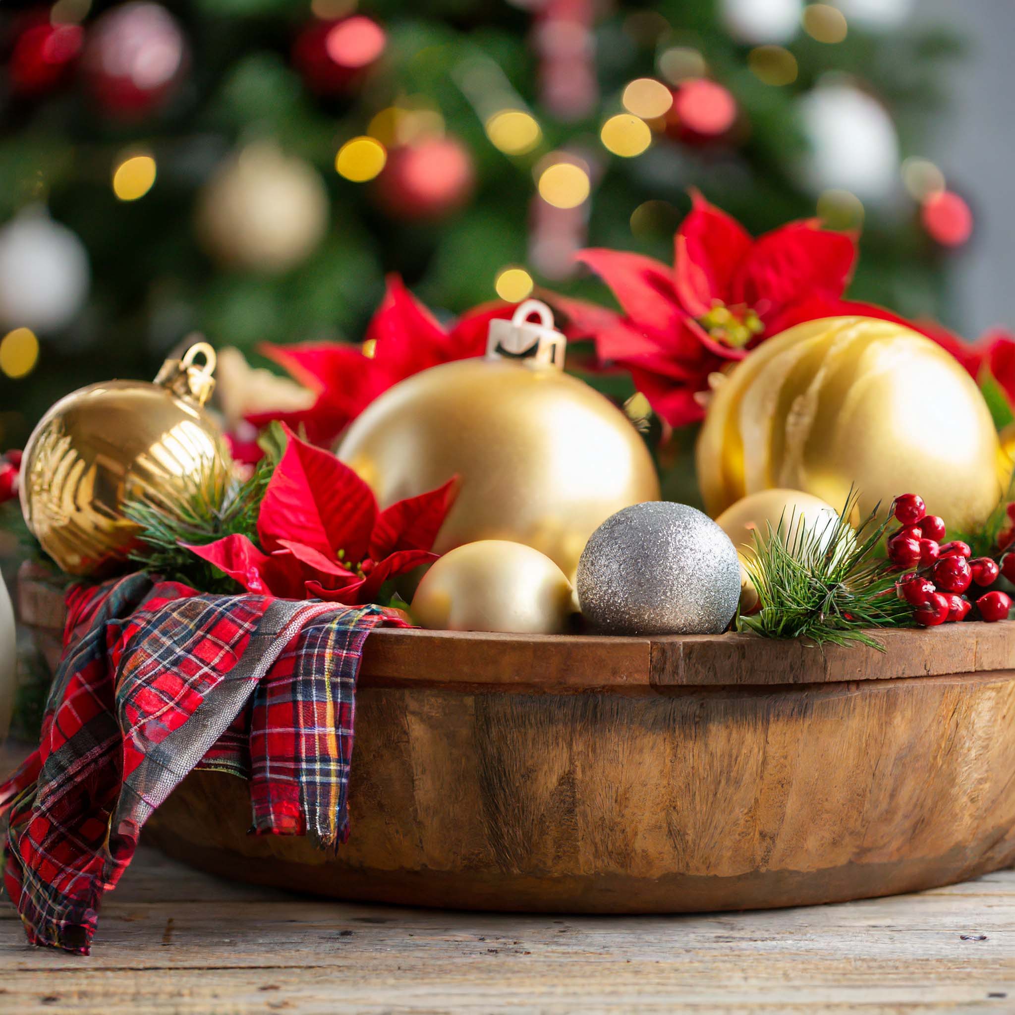 Wooden dough bowl filled with holiday ornaments, plaid ribbon, poinsettias, and berry sprays.