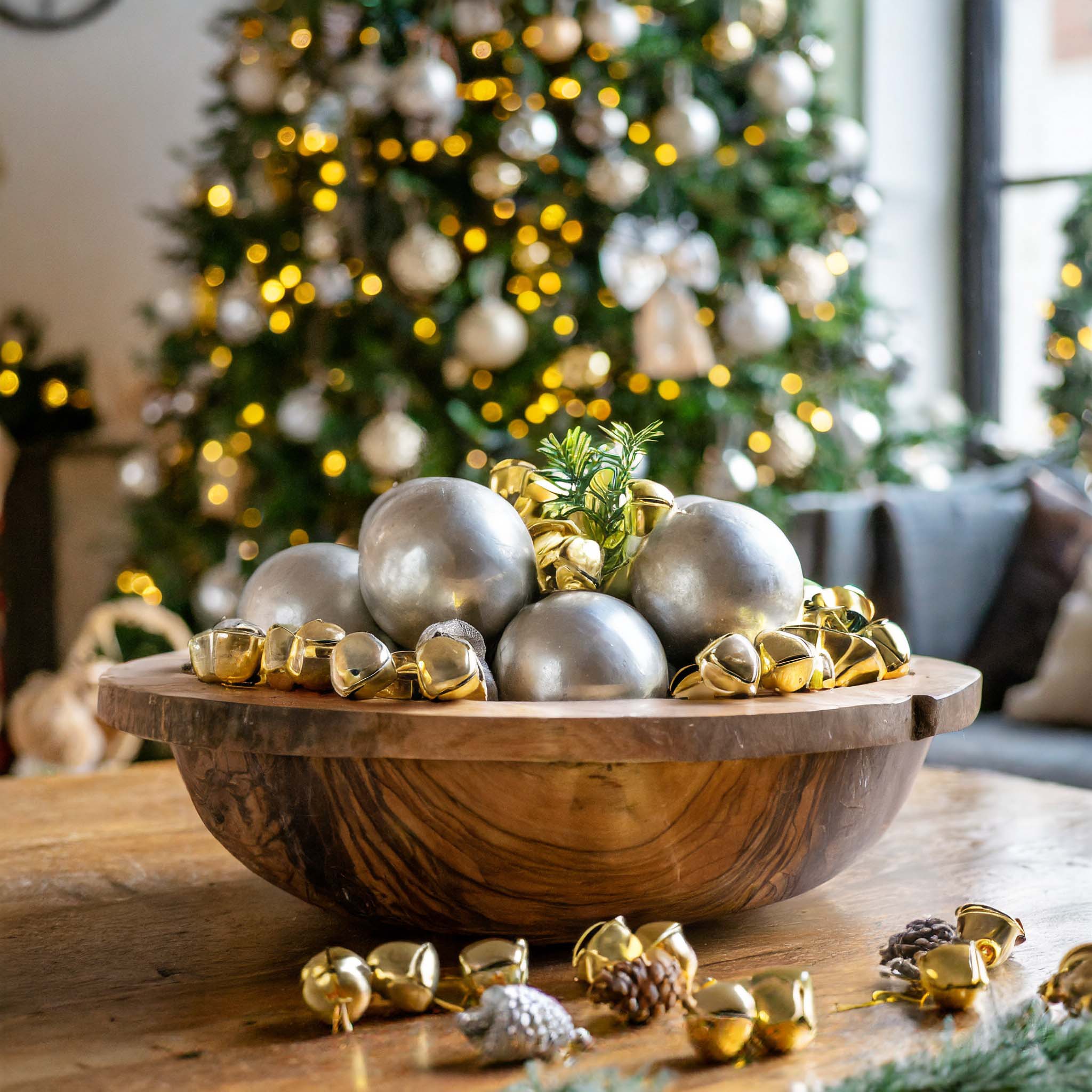 Wooden dough bowl filled with jingle bells and silver ornaments with Christmas tree in background.