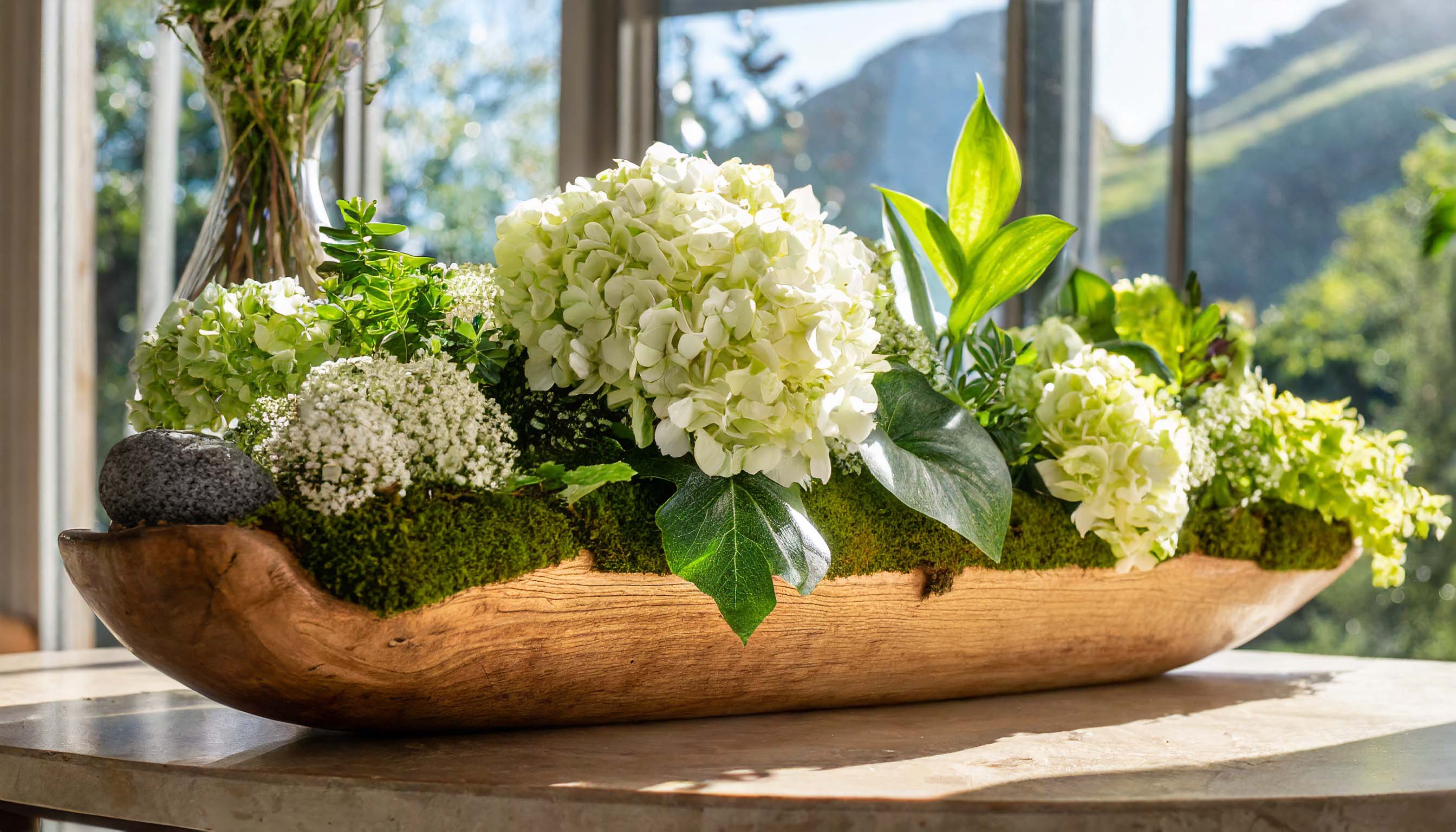 Wooden dough bowl filled with hydrangeas, plants, and moss in front of sunny window.