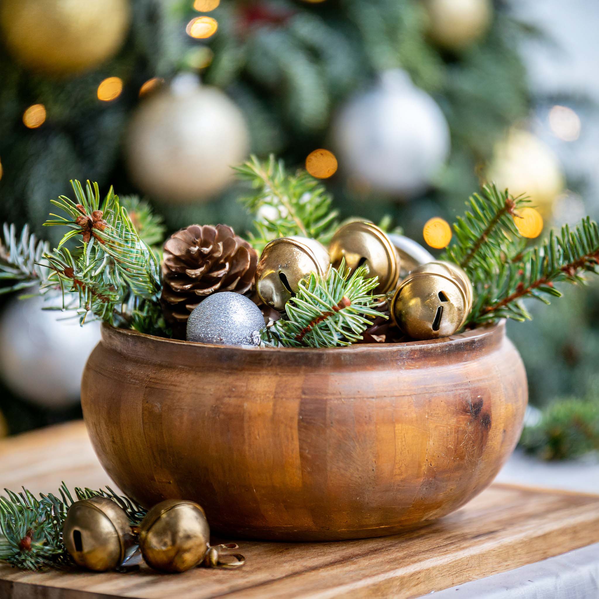 Wooden dough bowl filled with brass jingle bells, ornaments, and pine branches.