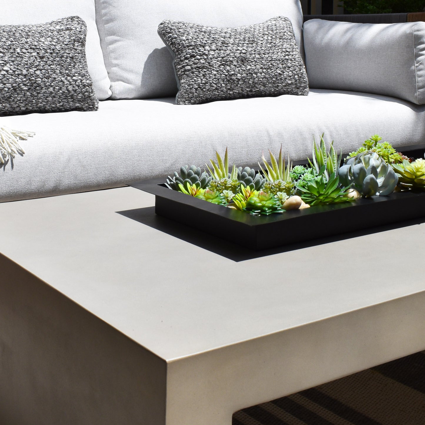 Black matte metal centerpiece tray filled with succulents on gray concrete table with light gray fabric outdoor sofa and dark gray large knot pillows.