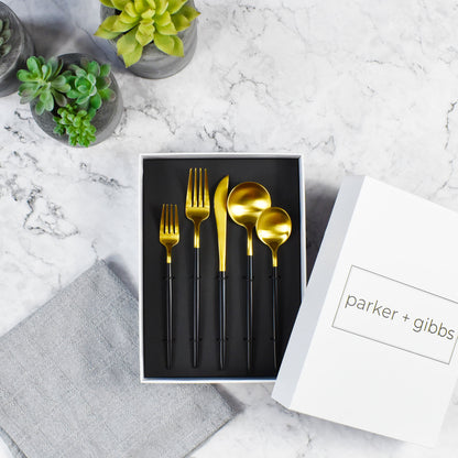 Brushed gold and black handle flatware in custom box on marble countertop with succulents in concrete planters.