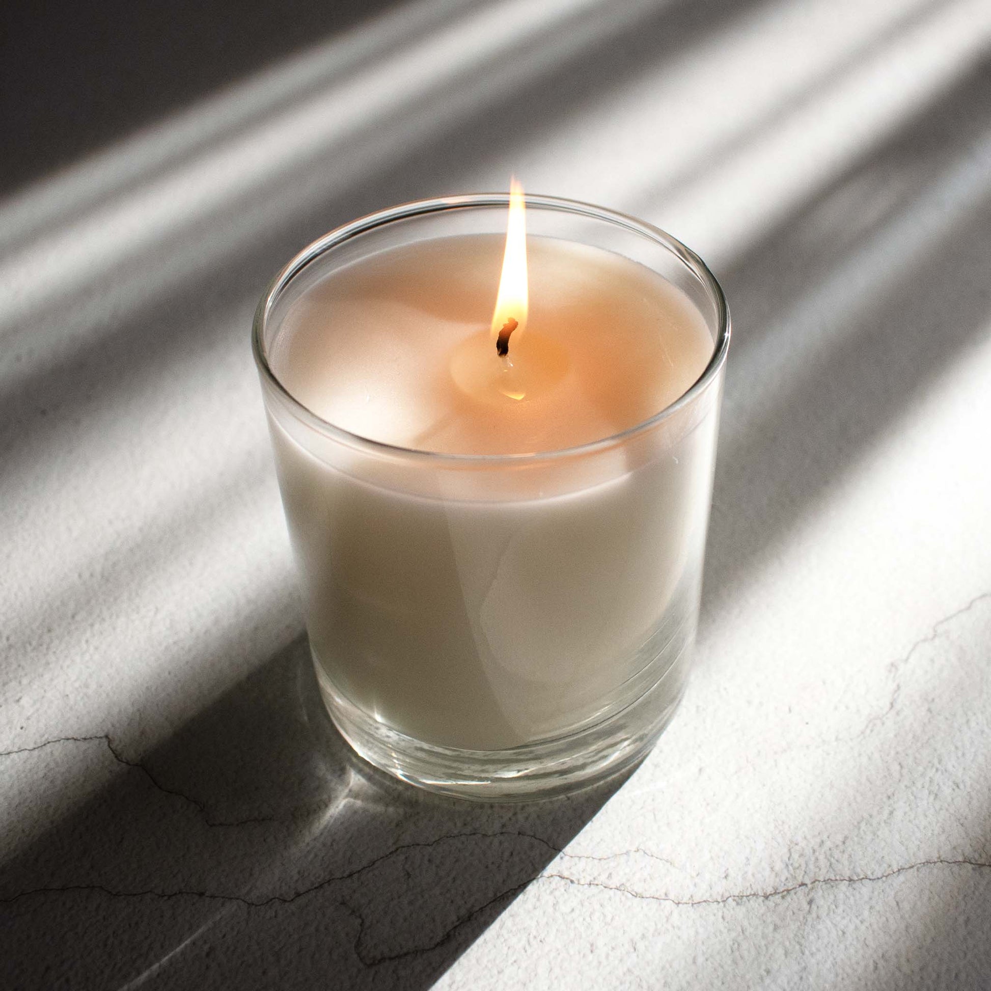 Burning scented soy blend wax candle on limestone table with sunrays.