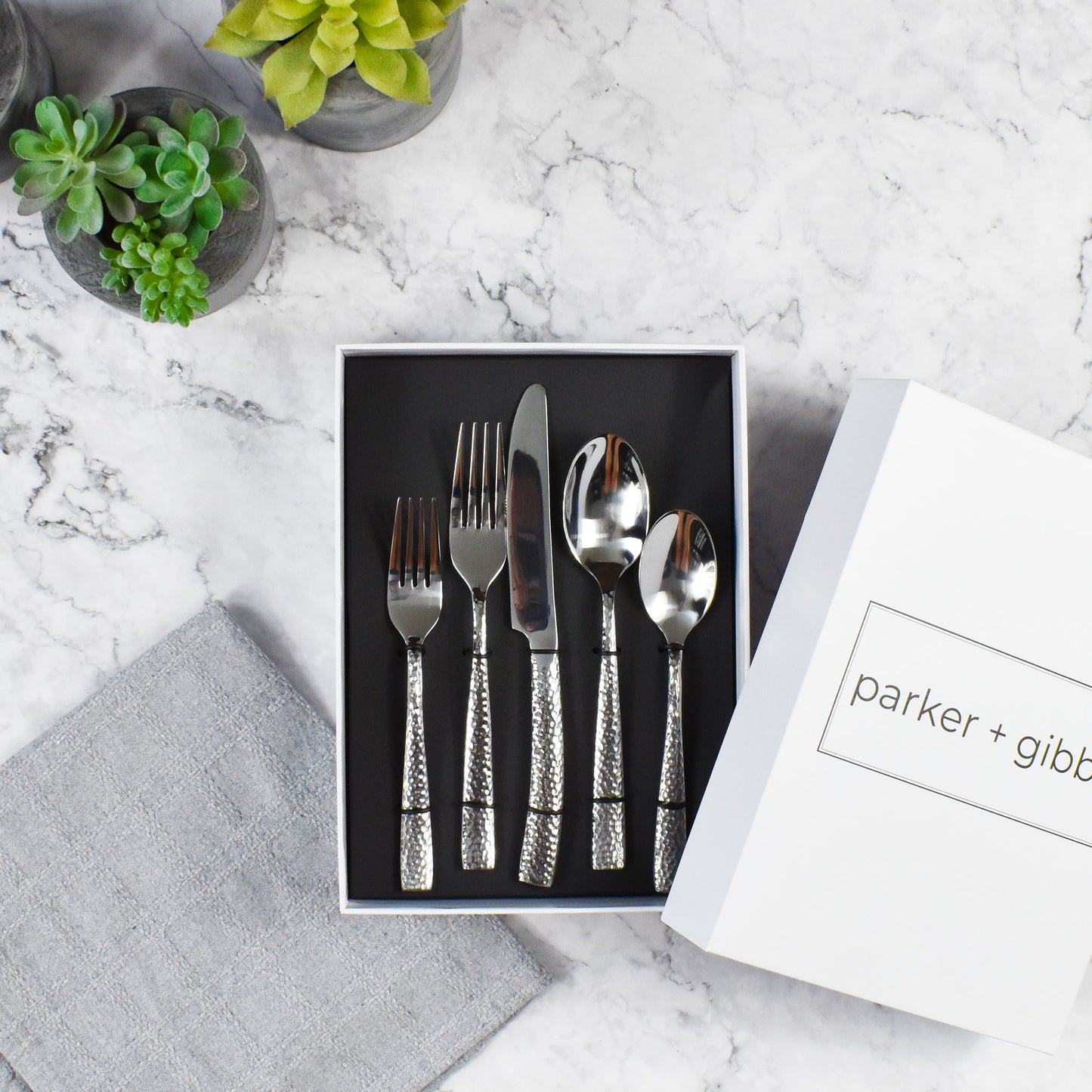 Polished silver hammered flatware in custom box on marble countertop with succulents in concrete planters.