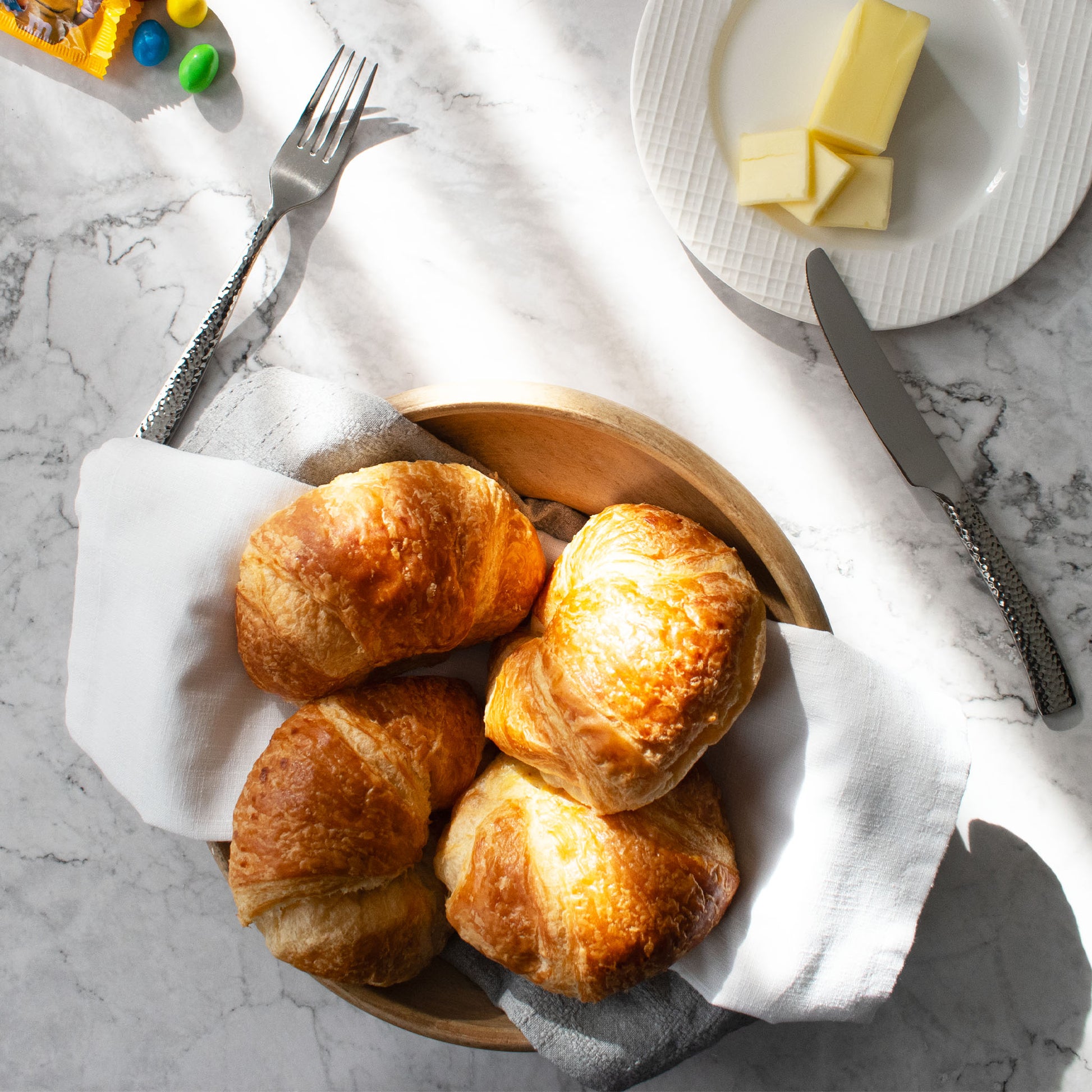 Polished silver hammered flatware on marble table with croissants and butter.