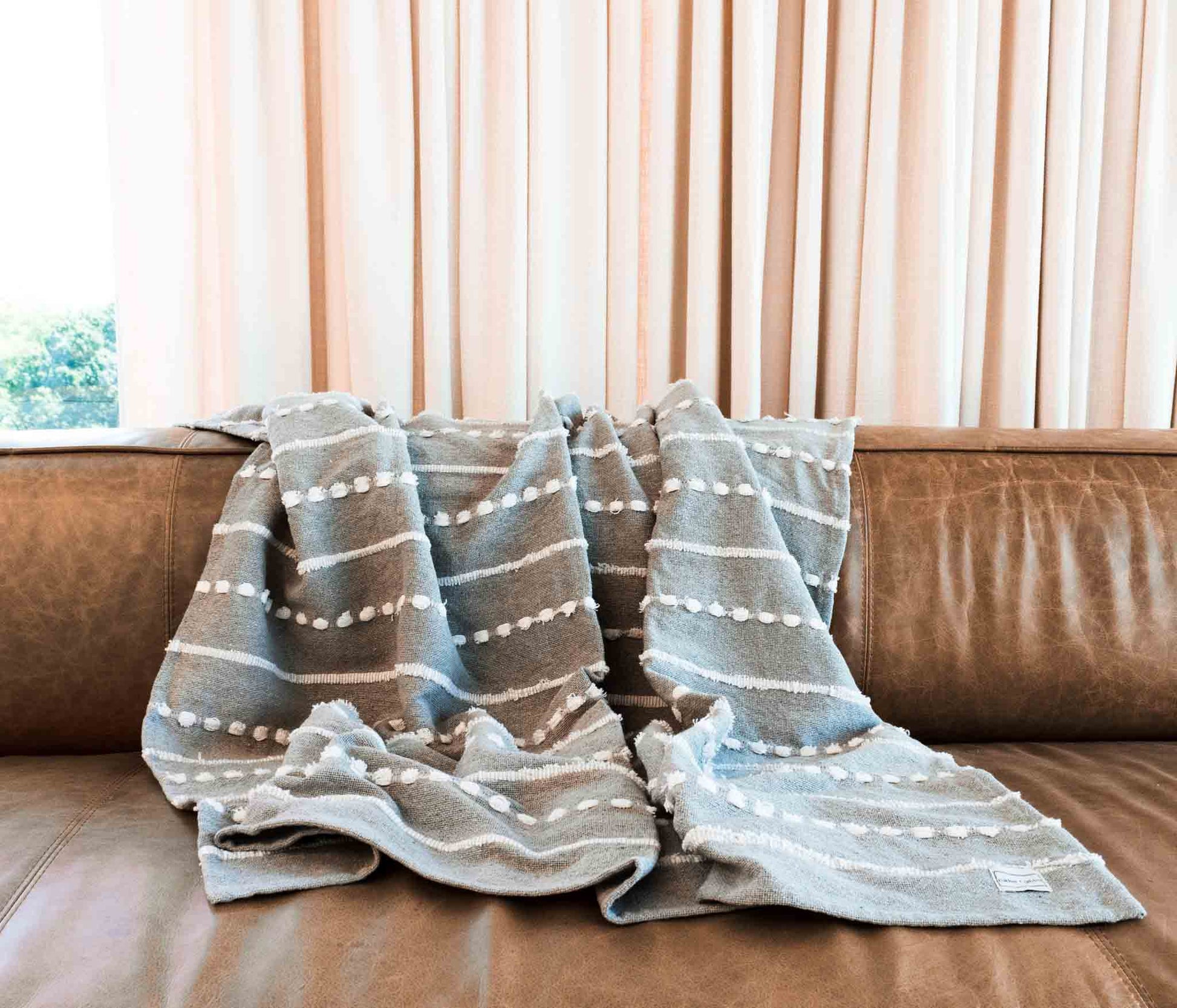 Cotton throw blanket with gray stitching and white tufted stitched stripes on leather sofa with drapery in background.