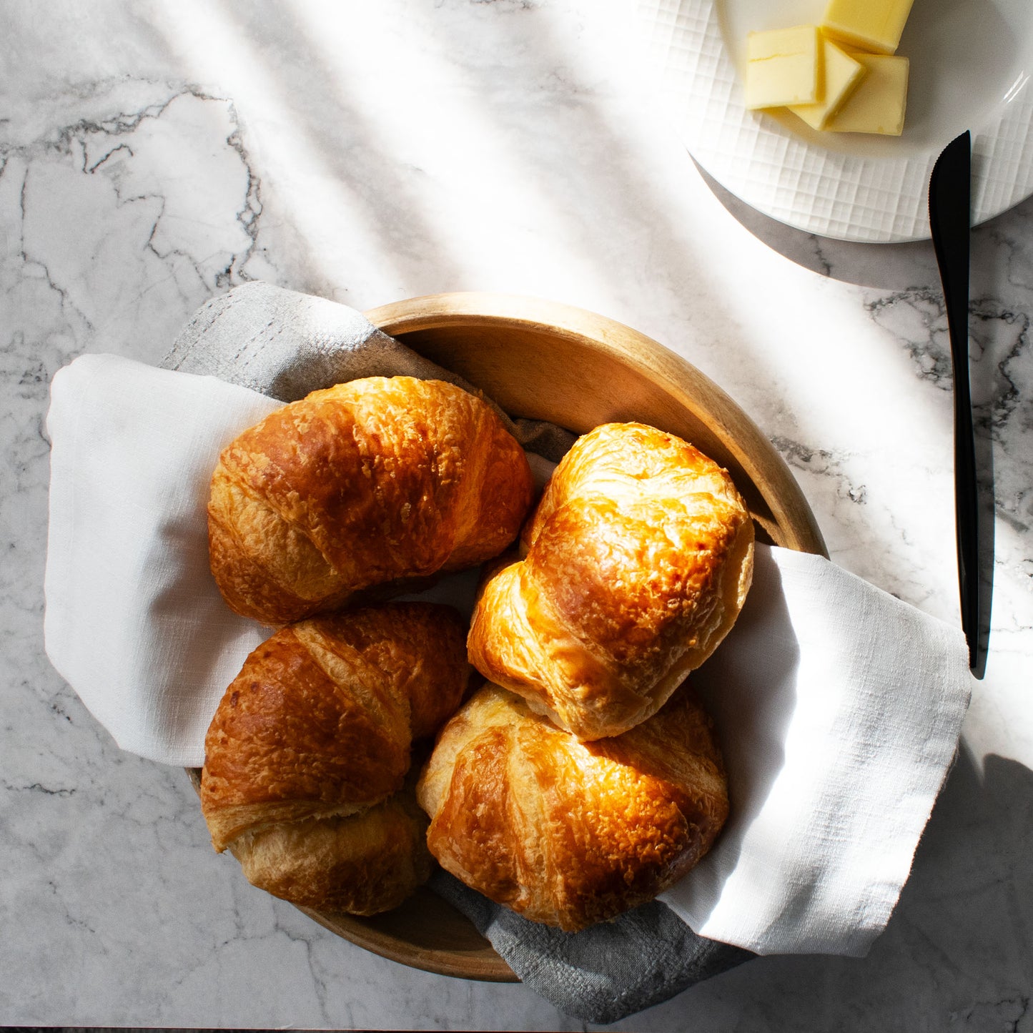 Mirrored polished black flatware on marble table with croissants and butter dish.