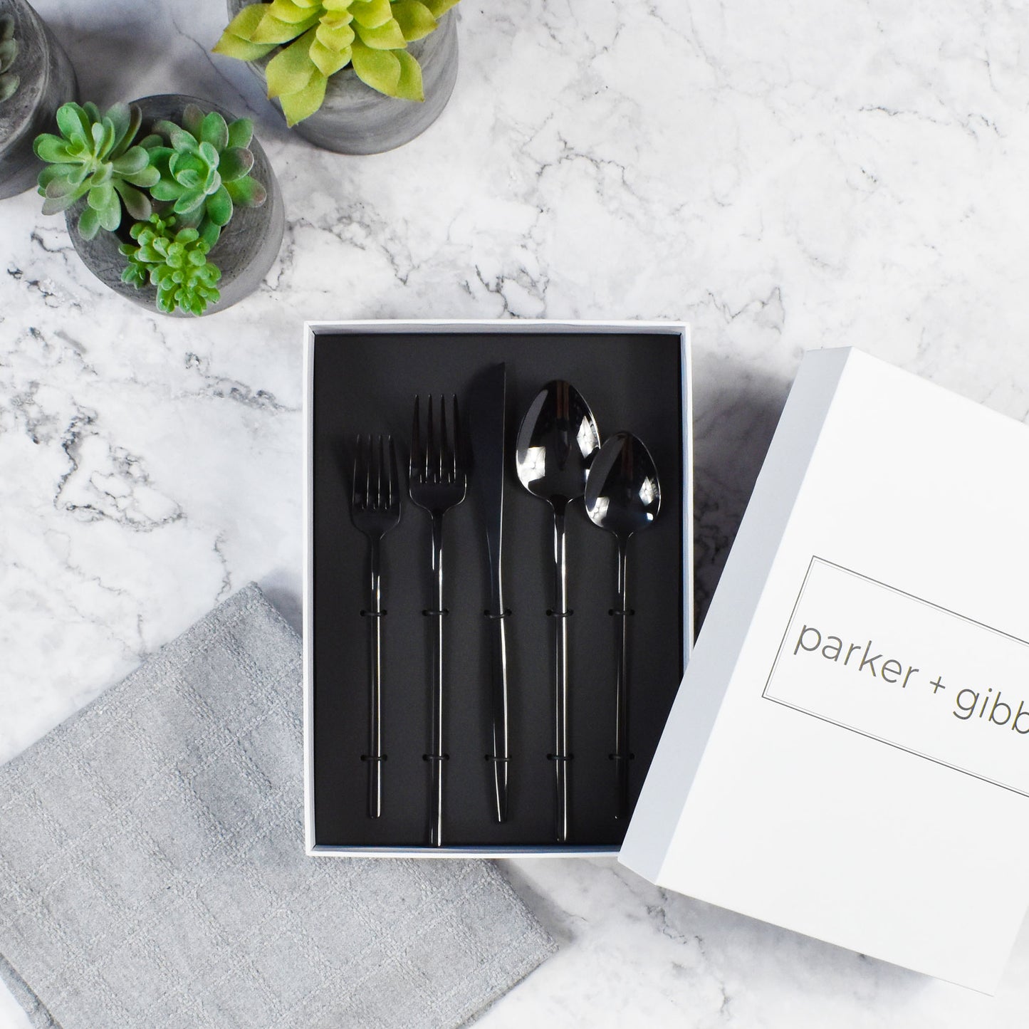 Mirrored polished black flatware in custom box on marble countertop with succulents in concrete planters.