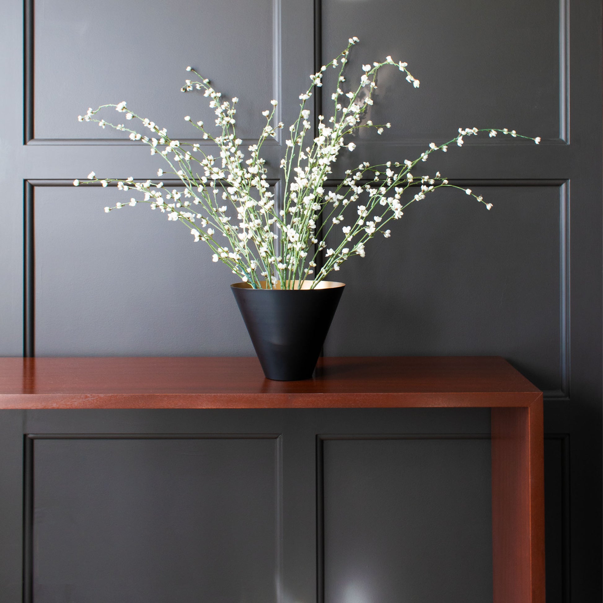 Black painted votive-shaped vessel with gold painted interior filled with cherry blossoms on modern mahogony console table in front of dark gray paneled wall.