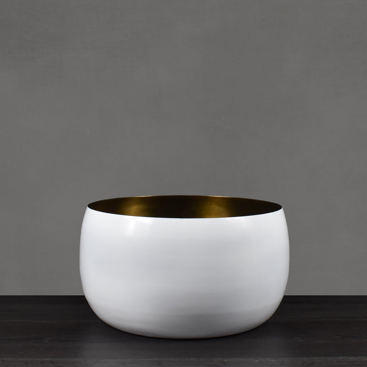 White enamel painted vessel with antiqued brass interior finish on dark wood floor with gray background.