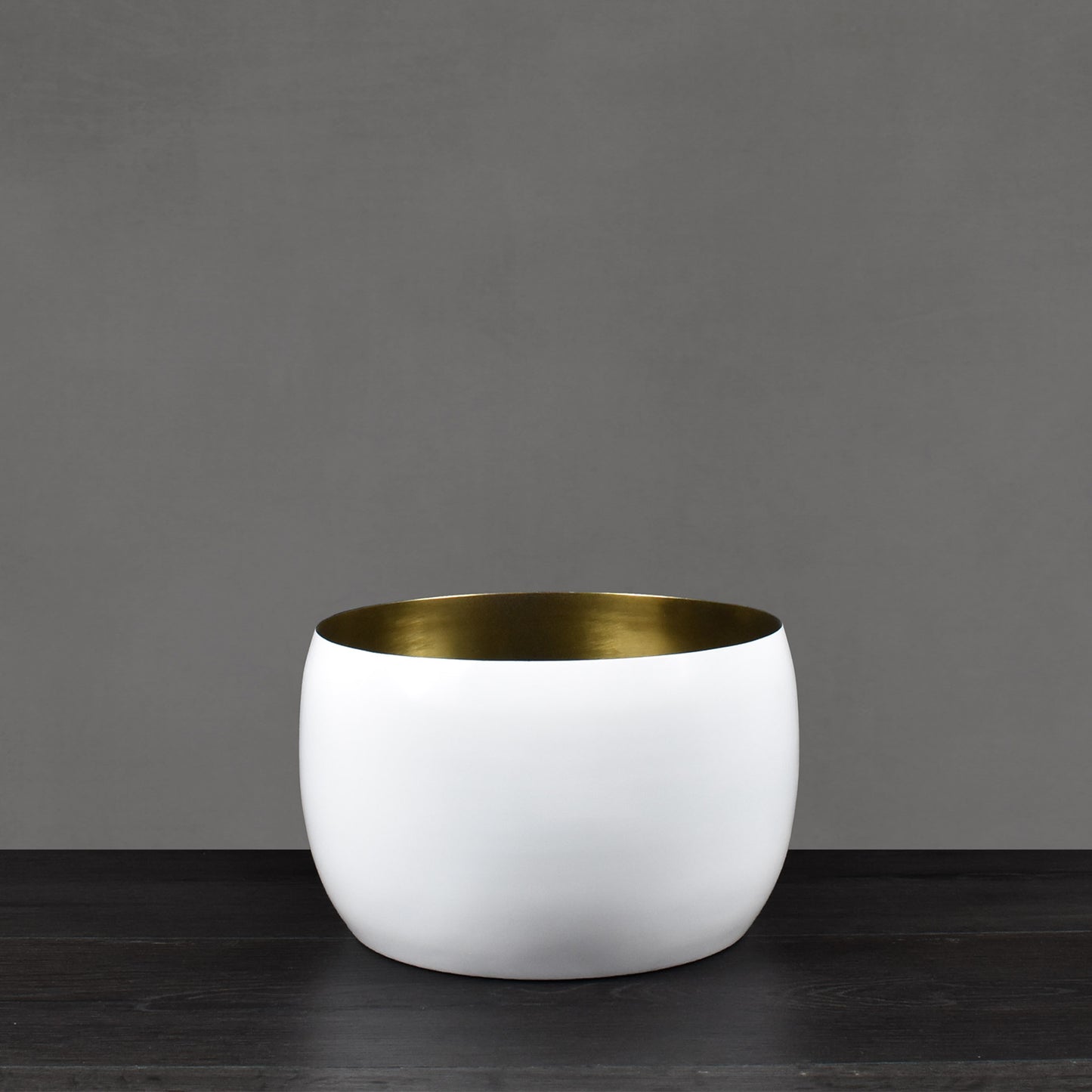 White enamel painted vessel with antiqued brass interior finish on dark wood floor with gray background.