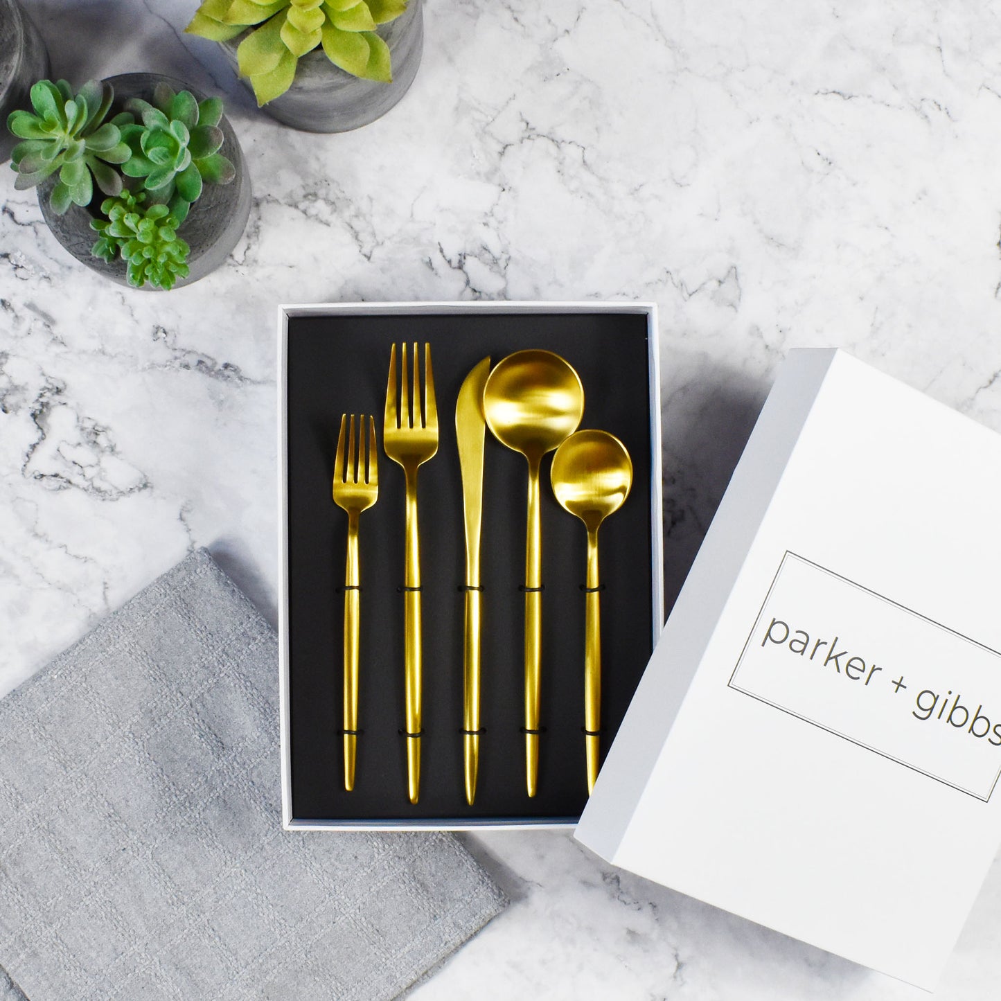 Matte brushed gold flatware in custom box on marble countertop with succulents in concrete planters.