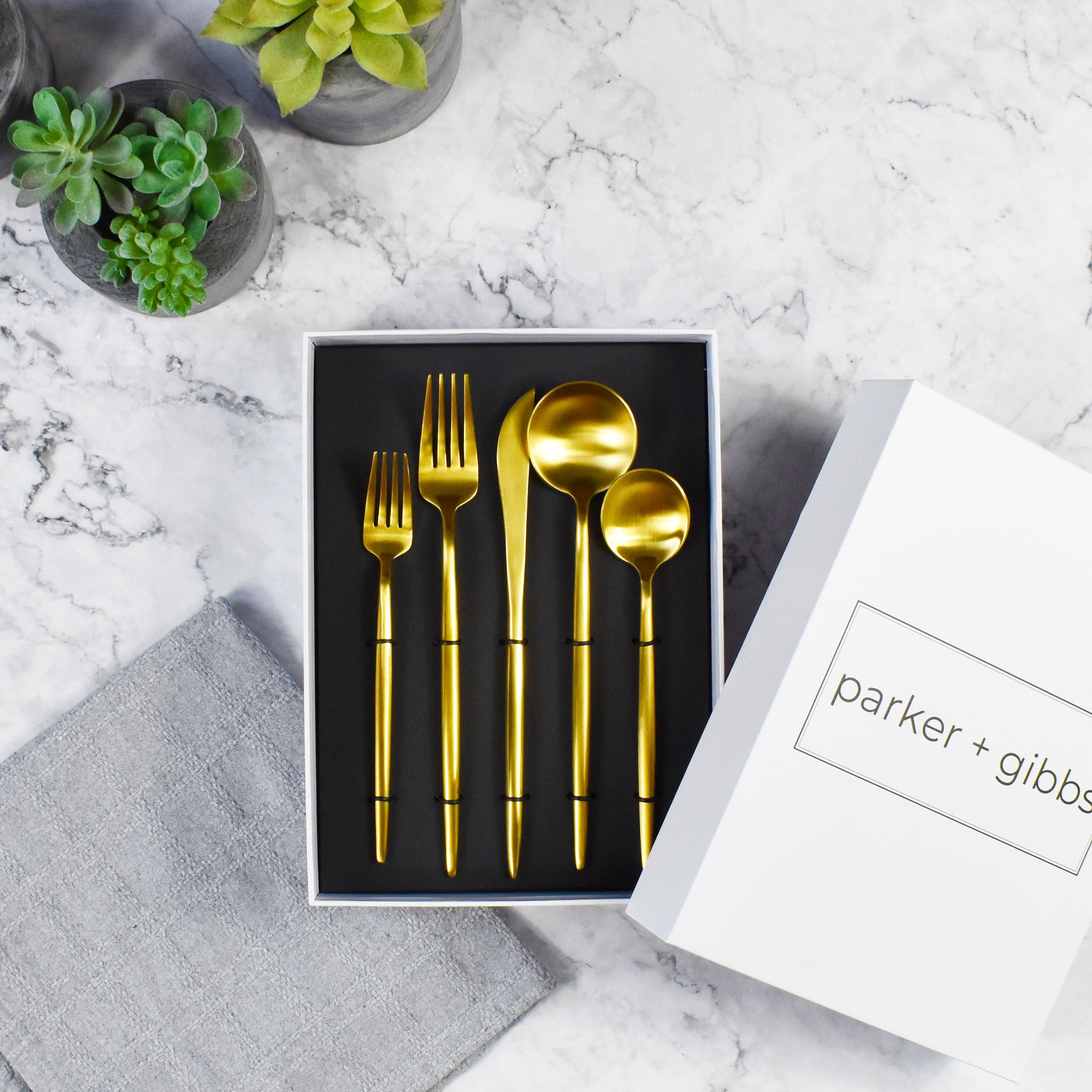 Matte brushed gold flatware in custom box on marble countertop with succulents in concrete planters.