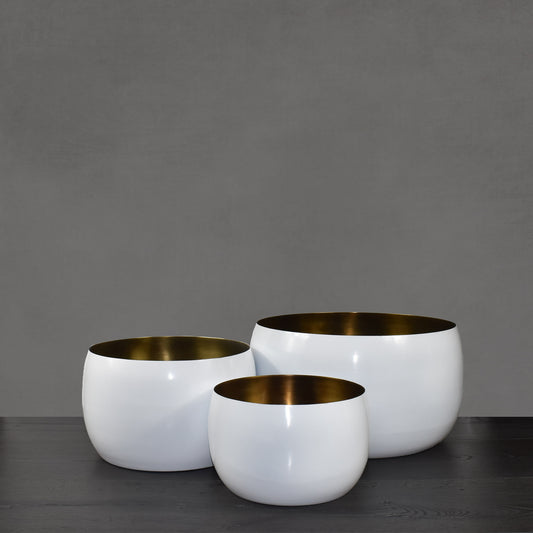 Trio of white enamel painted vessels with antiqued brass interior finish on dark wood floor with gray background.