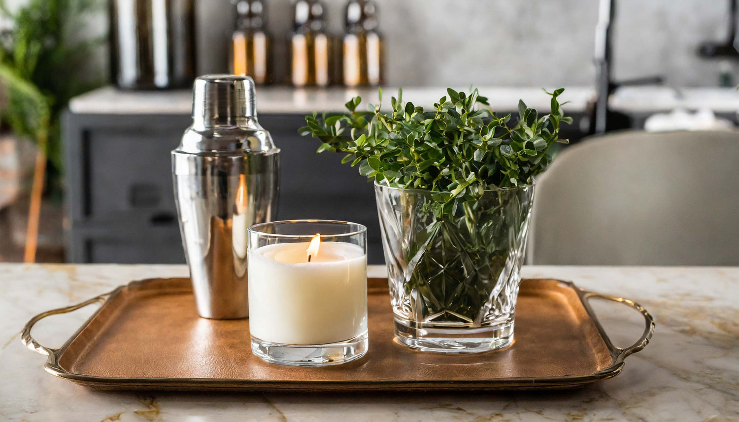 Scented soy candle on leather tray, cocktail shaker, greenery in vase.