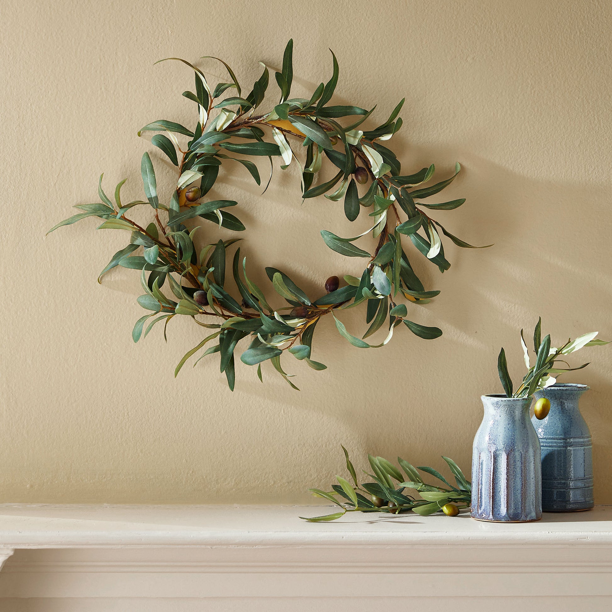 16" faux olive wreath hanging above mantel with pottery vases.