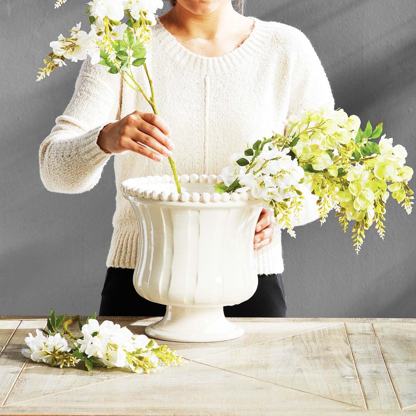 Woman filling a beaded white flared ceramic vase with floral stems.