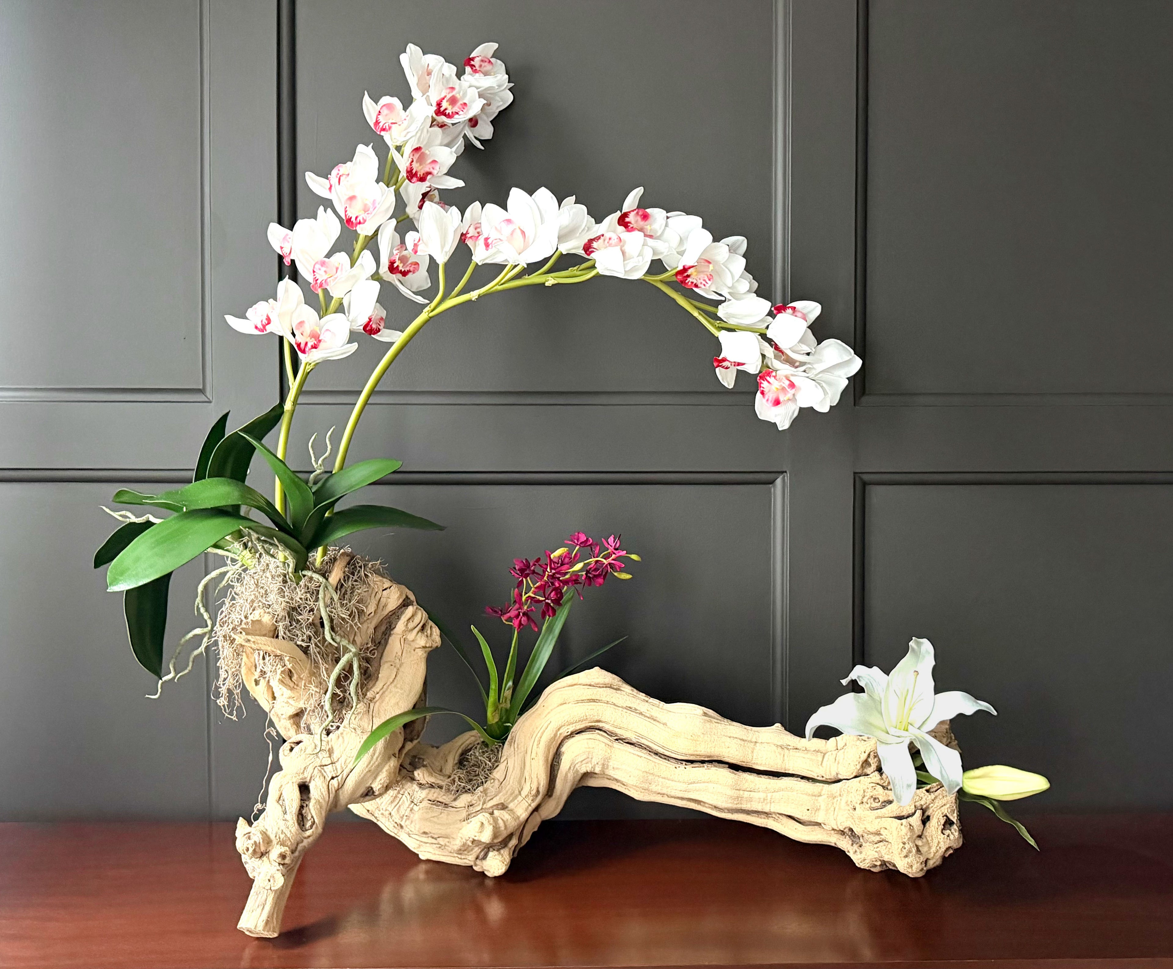 Decorative floral arrangement with white and pink flowers on a wooden base against a dark panel wall.