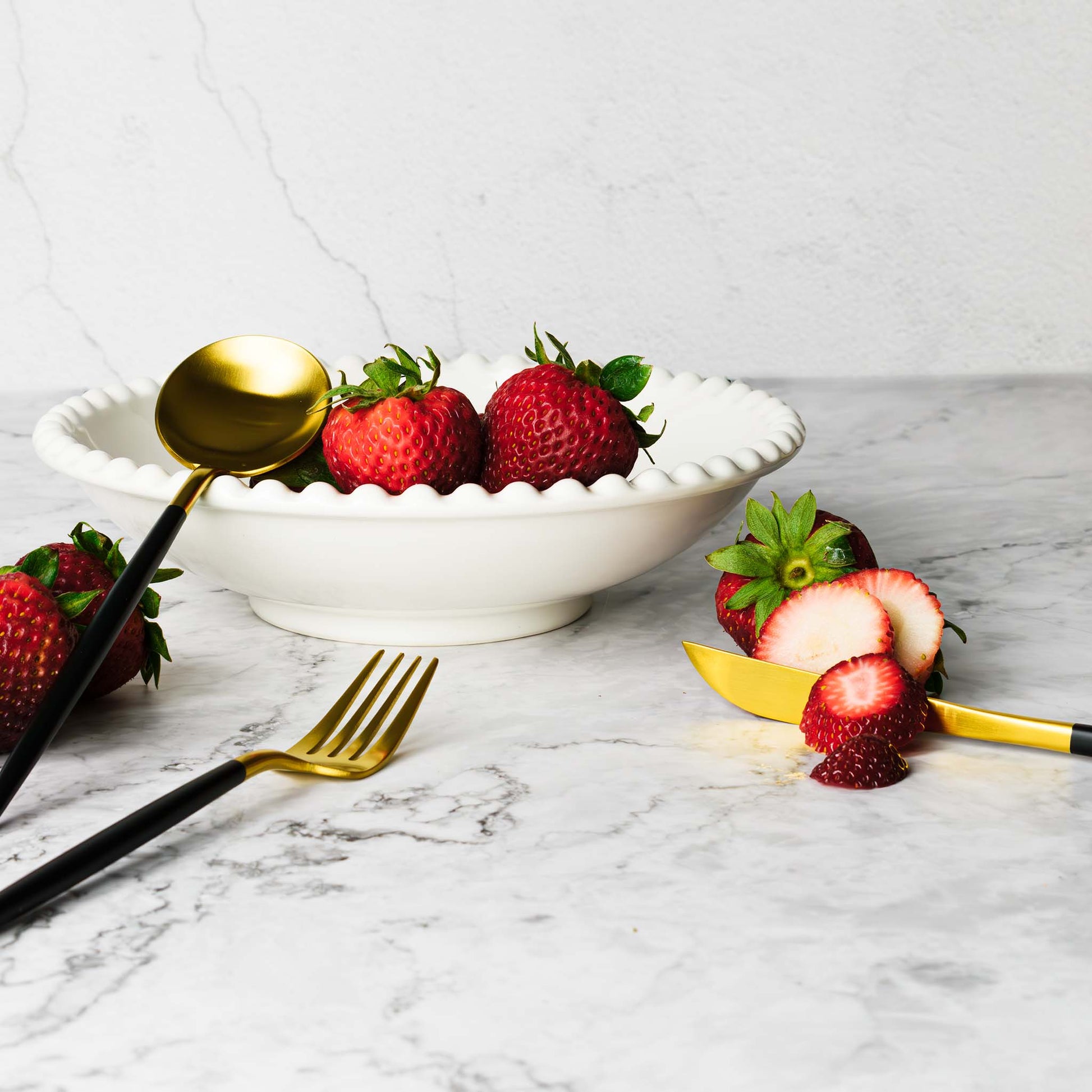 Flatware set in brushed gold and black, with strawberries in bowl, on marble table.