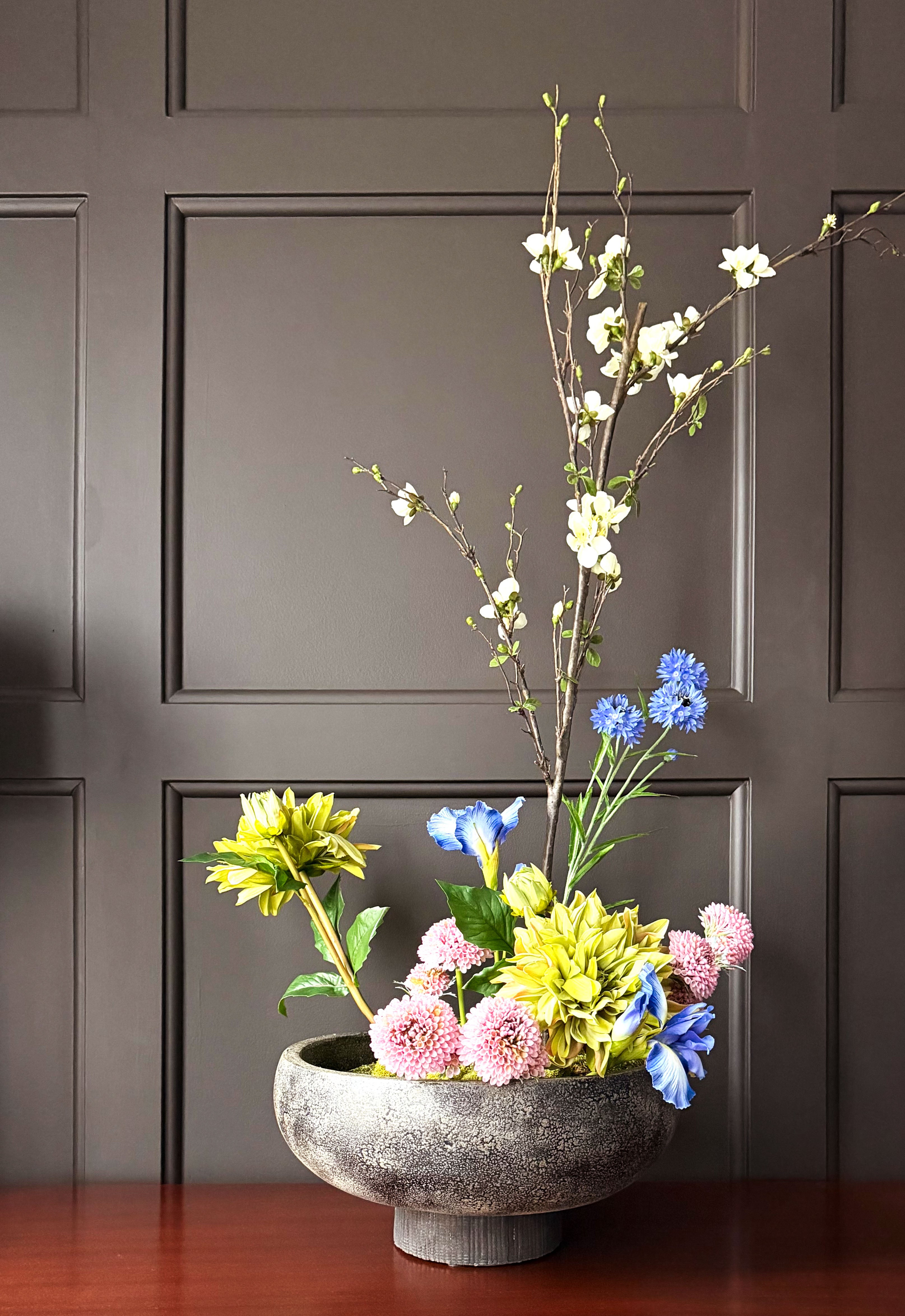 Decorative flower arrangement in a stone bowl against a dark paneled wall.
