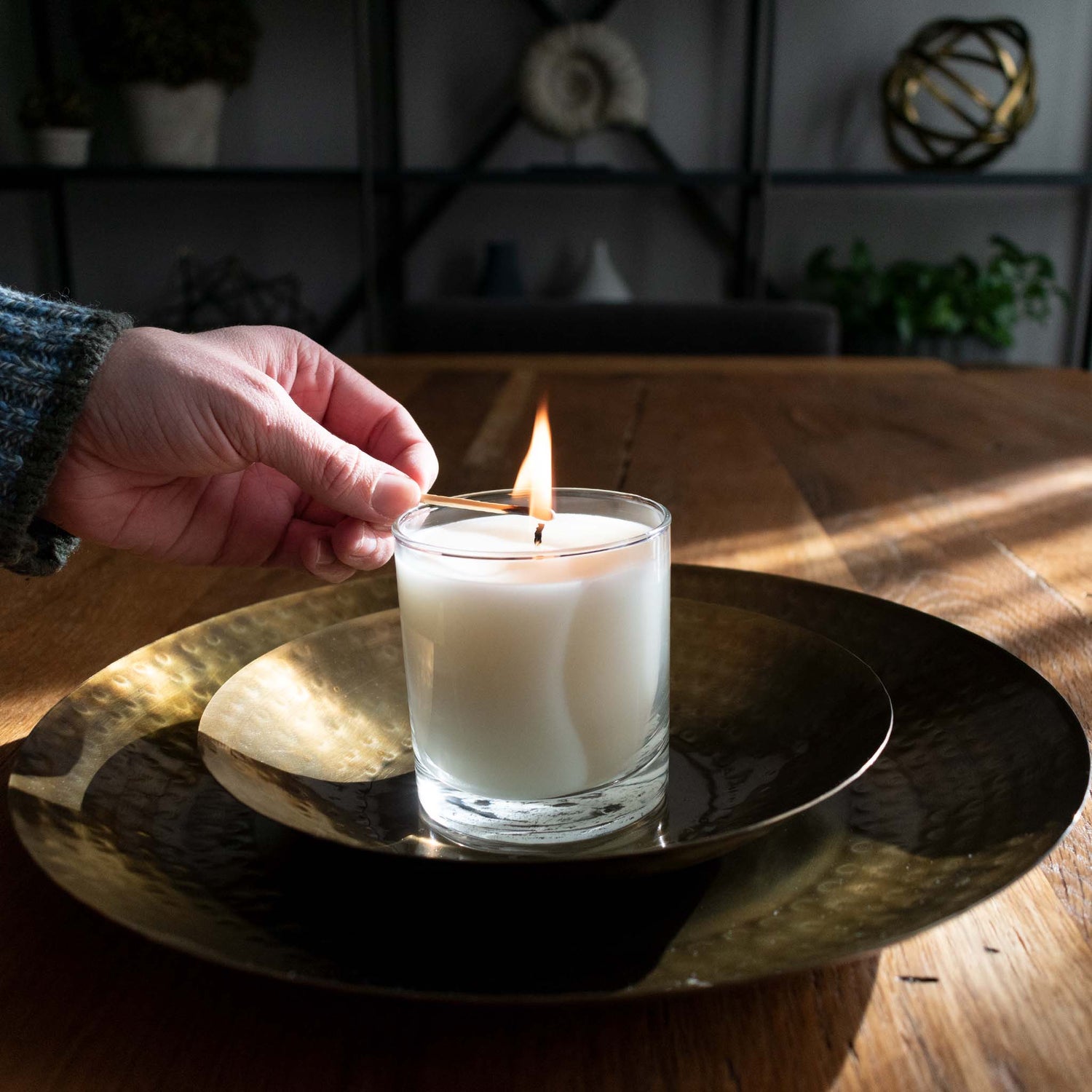 Person lighting white soy wax candle on hammered brass trays on farmhouse table.