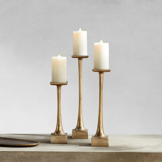 Modern pillar candleholders, small, medium and large, with champagne finish on table with gray background.