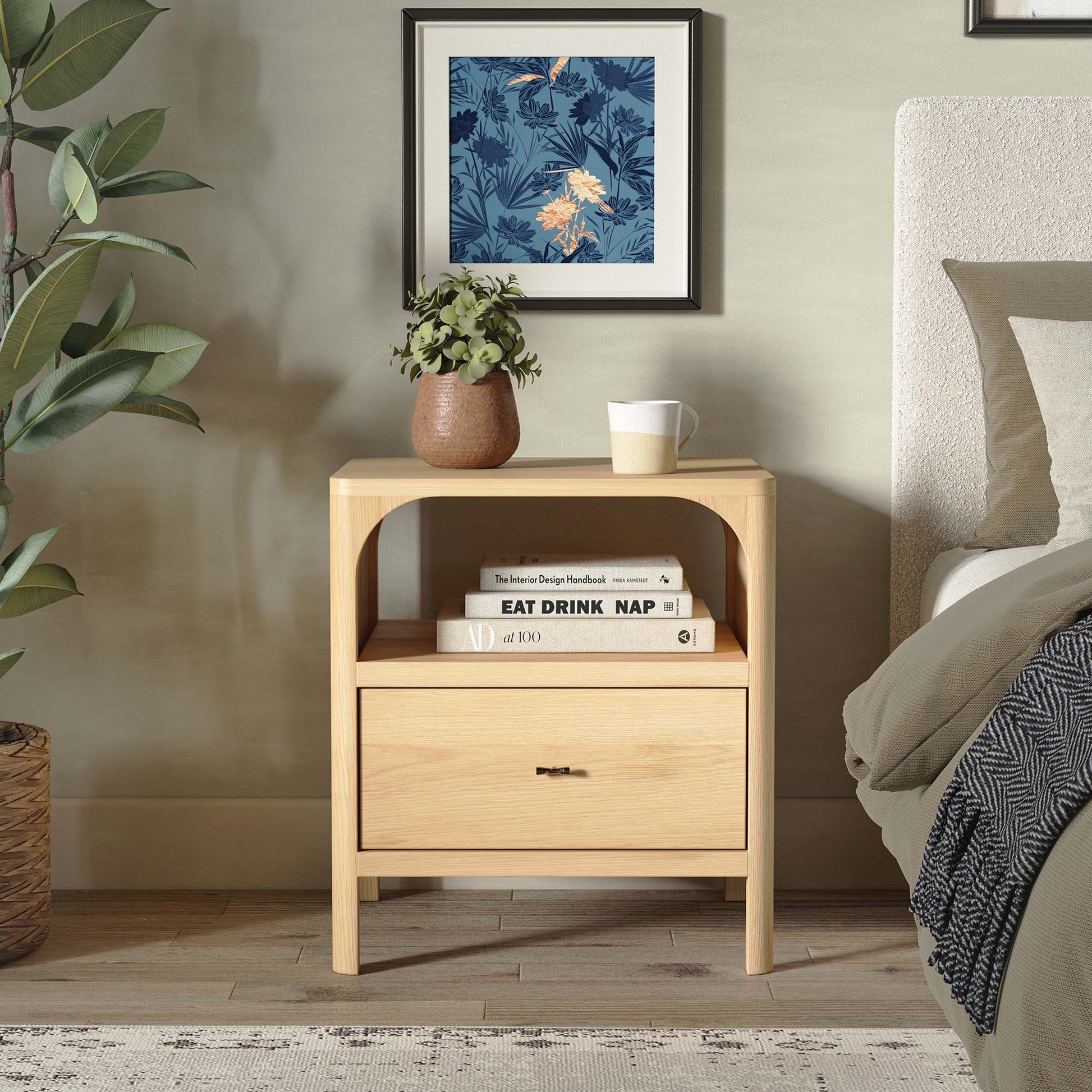 Wooden nightstand with books and a mug next to a bed with a floral painting on the wall.