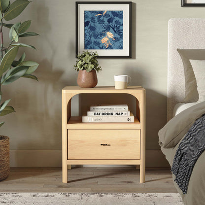Wooden nightstand with books and a mug next to a bed with a floral painting on the wall.