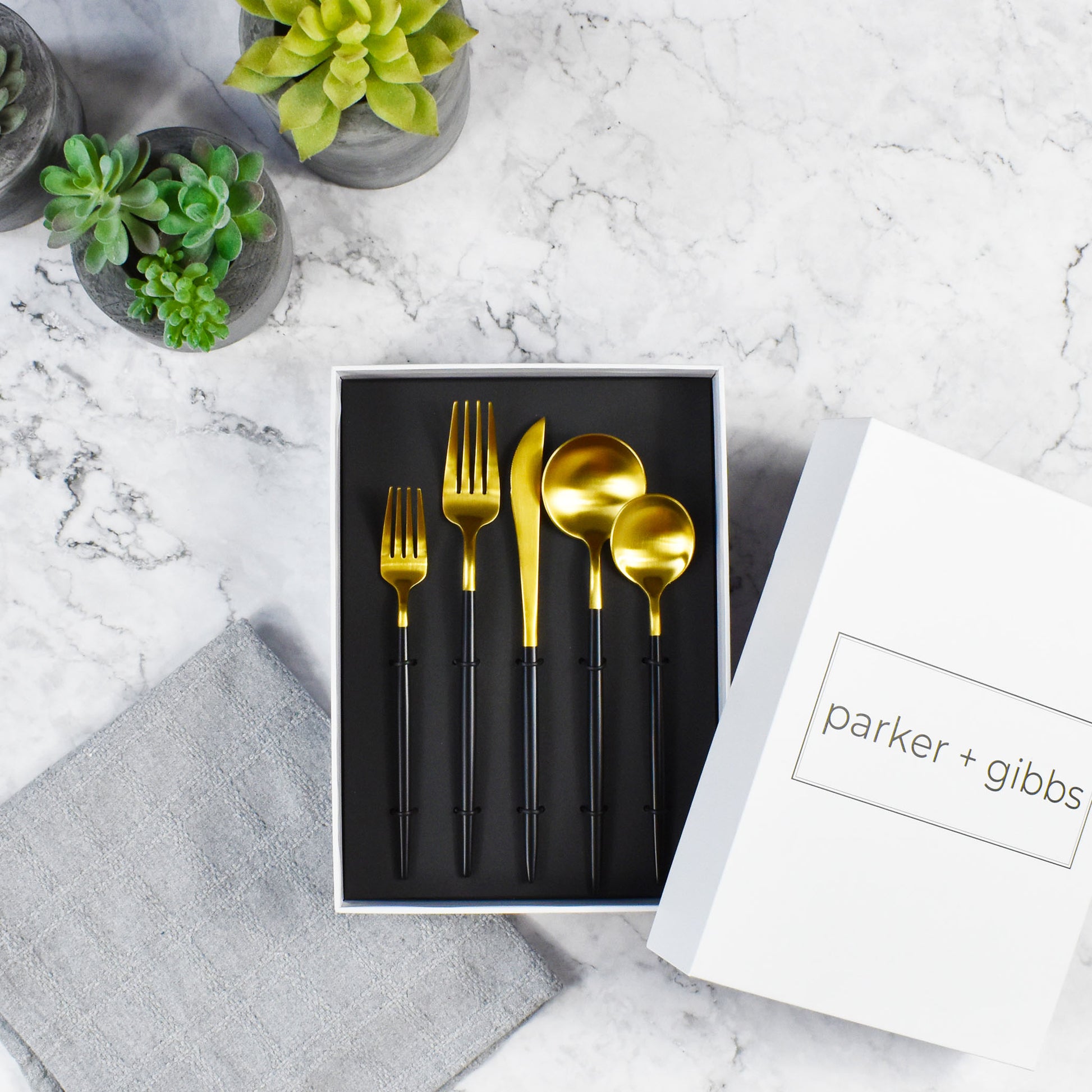 Brushed gold and black handle flatware in custom box on marble countertop with succulents in concrete planters.